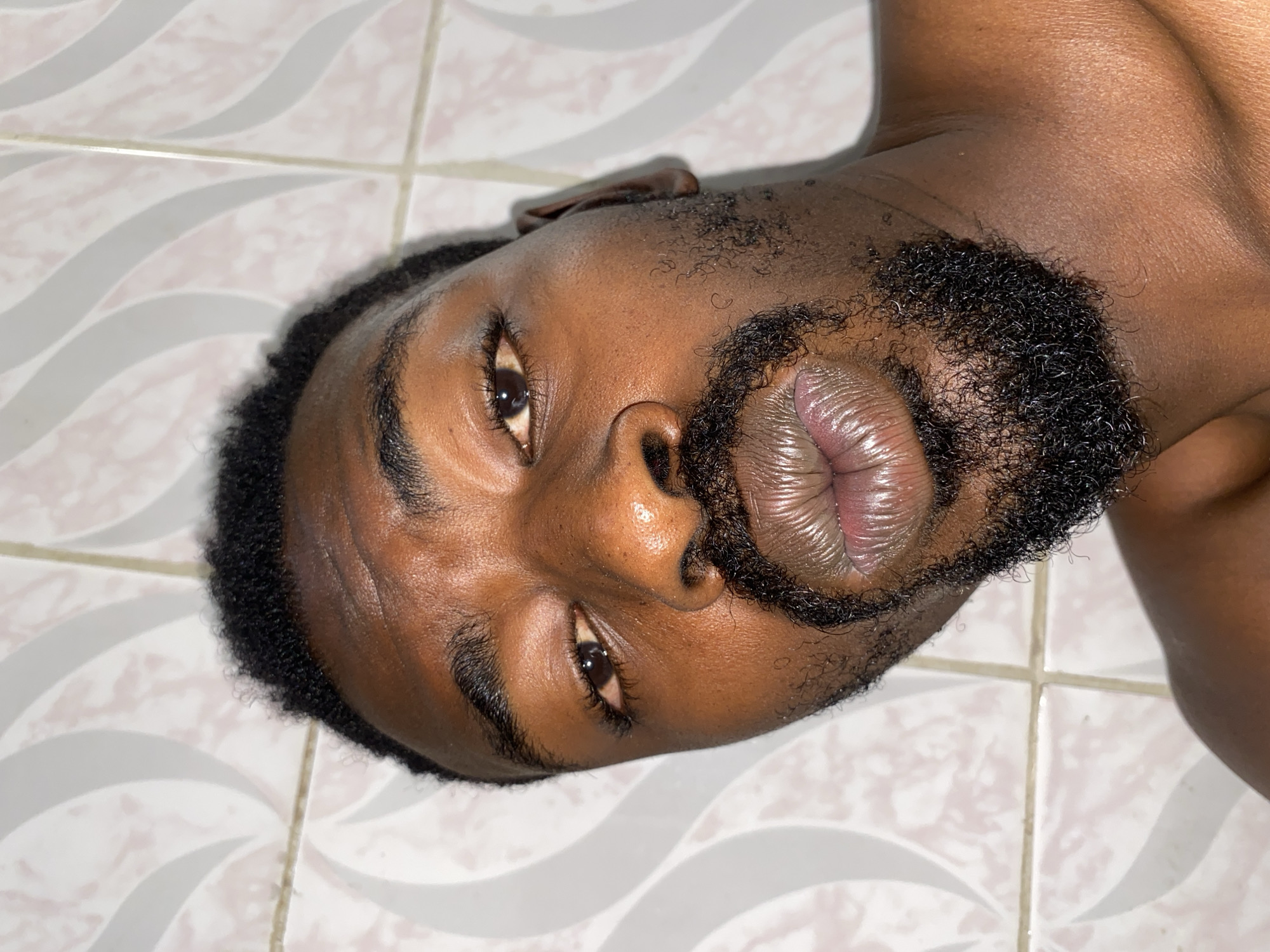 An overhead, close-up portrait shot depicts the head and upper chest of a dark-skinned individual, rotated 90 degrees clockwise. The subject, likely male, has short, tightly curled dark hair and a neatly trimmed dark beard that covers the chin and jawline. Their gaze is directed straight towards the camera, with dark eyes and well-defined eyebrows. The nose is prominent, and the full lips appear glossy and slightly parted or pursed, revealing fine textural lines. The skin on the face, neck, and visible shoulder area is smooth and reflects light, suggesting moisture or an oily sheen.
The individual is lying on a light-colored tiled floor, which serves as the background. The tiles feature a repeating pattern of subtle, wavy lines in a lighter shade, possibly white or light gray, against a neutral-toned base, with visible grout lines separating them. The lighting appears to be an indoor setting, casting highlights on the subject's features. The overall impression is a direct and focused portrait, emphasizing facial details and skin textures. No specific activity is taking place other than posing. There is no visible text in the image.