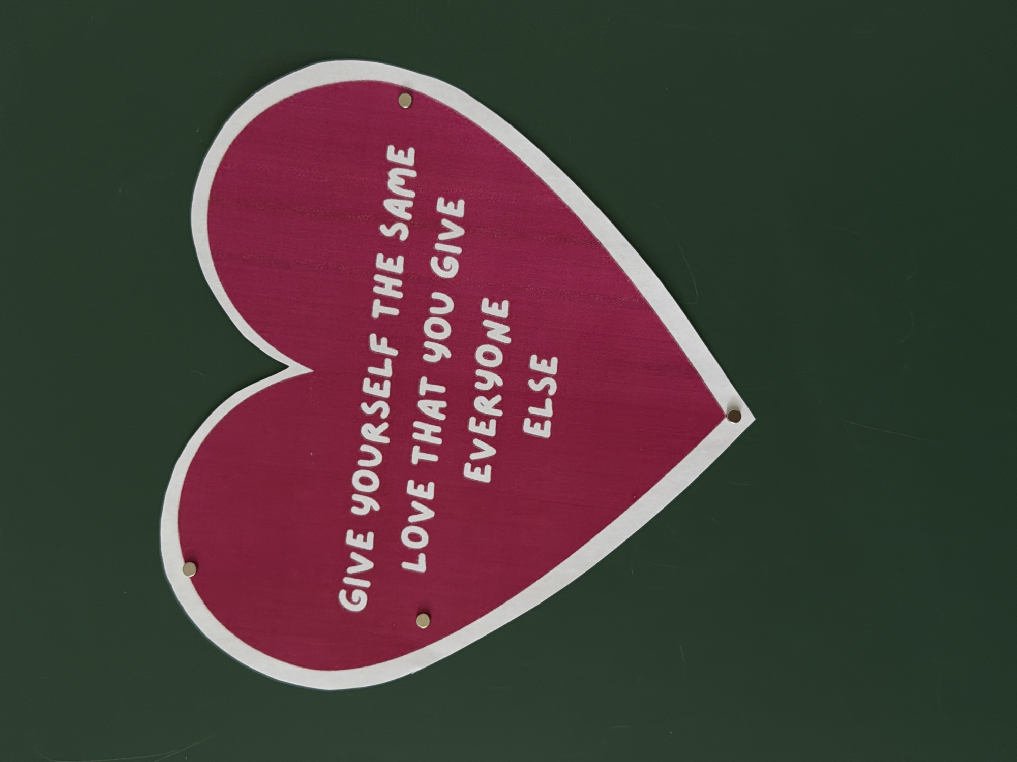 A rotated, medium shot shows a wooden, heart-shaped sign with the words "GIVE YOURSELF THE SAME LOVE THAT YOU GIVE EVERYONE ELSE" painted in white on a burgundy background. The heart shape is outlined in white and is attached to a dark green wall with three small, round, brass-colored tacks. The environment suggests an indoor setting, possibly a shop, home, or community space. The location is unknown.