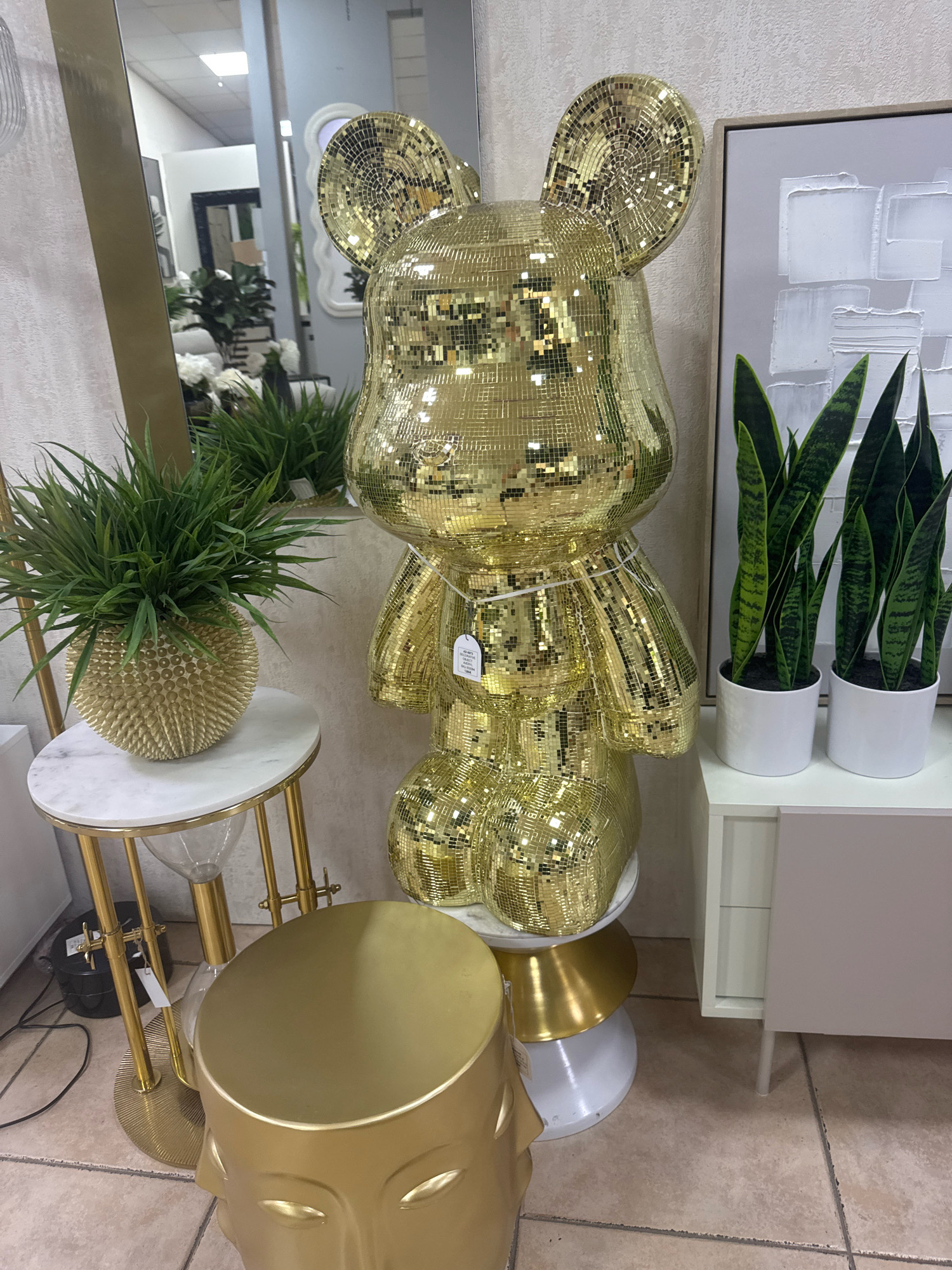 An interior scene at a retail establishment in Agios Athanasios Municipality, Cyprus displays a large, gold, sequined teddy bear statue as the central focus. The statue is positioned on a white and gold pedestal, with its surface covered in small, reflective gold squares. To the left of the statue is a small, oval marble-topped side table with gold legs, holding a spiky gold vase filled with green artificial grass. Further left, a tall, thin gold floor lamp is partially visible. To the right of the statue, two white pots containing snake plants sit on top of a light grey cabinet. Behind the plants and cabinet, a framed abstract artwork with white textured squares on a grey background is mounted on the wall. A large mirror on the left side of the image reflects more of the store's interior, including other furniture and decorative items. The floor is tiled in a neutral, earthy tone. No people are present in the frame.