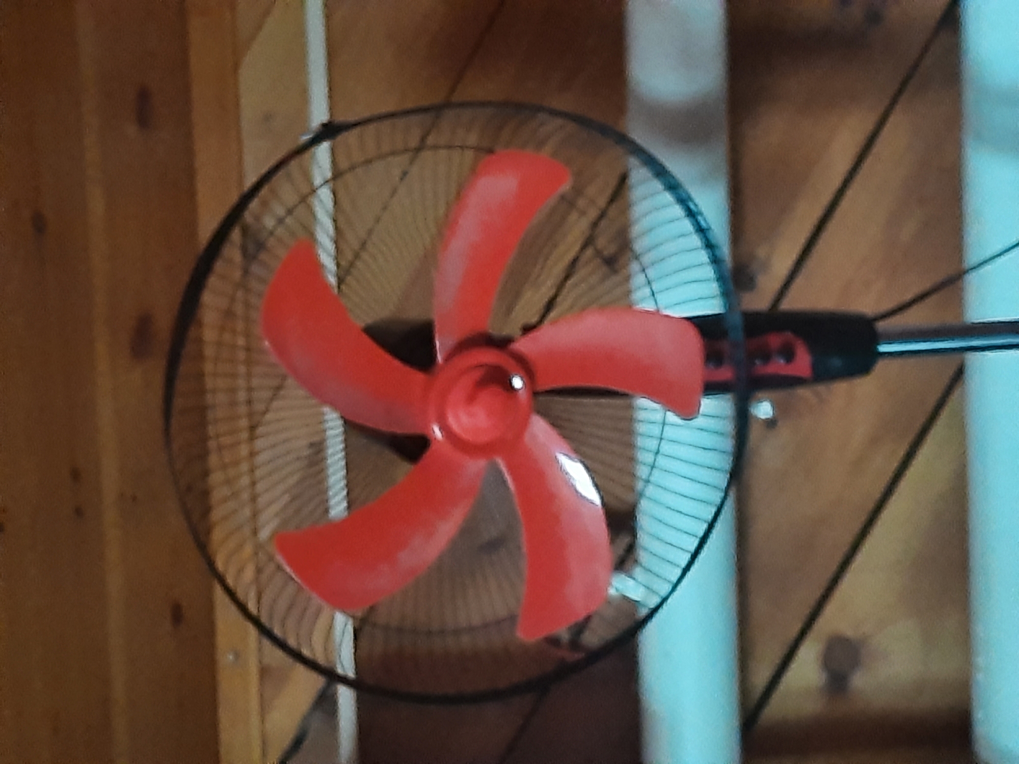 The image shows a close-up, eye-level shot of a red oscillating fan. The fan has black blades and a black protective grill. It is positioned in front of a wooden wall. The fan is the primary subject, and its vibrant red color stands out against the darker background. There is no indication of people or any specific activity taking place, aside from the fan's stationary position. The overall environment appears to be indoors, possibly a room or a workshop. The location of Antonovka, Russia, is not discernible from the visual information provided in the image.