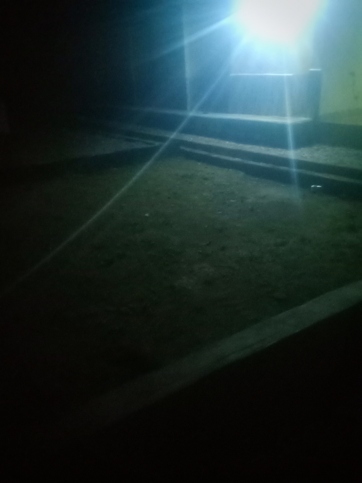 The image depicts an outdoor scene at night, illuminated by a bright light source that causes lens flare and washes out details in its vicinity. In the background, a series of concrete steps lead up to a raised platform, suggesting a public space like a park or an amphitheater. The ground in the foreground appears to be rough terrain, possibly dirt or gravel, with some scattered debris. The overall atmosphere is dark and somewhat eerie due to the low light conditions and the stark contrast from the intense light. The location is identified as the Jalingo City stadium in Jalingo, Nigeria. No people or specific actions are visible in the image.