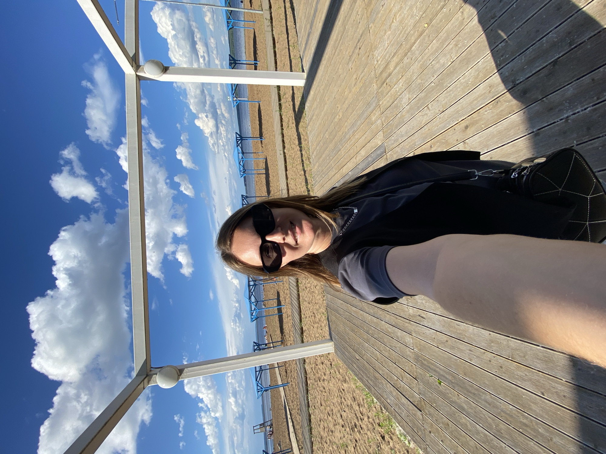 The image is a selfie taken by a woman on a wooden deck in front of a beach. She is wearing sunglasses, a dark vest over a grey t-shirt, and a necklace. She is holding the camera with her right arm extended. Behind her is a sandy beach with several blue beach umbrellas and some people lounging. The sea is visible in the background, under a bright blue sky with white clouds. The scene appears to be located at a beach resort or recreational area in the city of Chaikovsky, Russia, likely on the Kama River reservoir. The overall lighting suggests it is a sunny day.