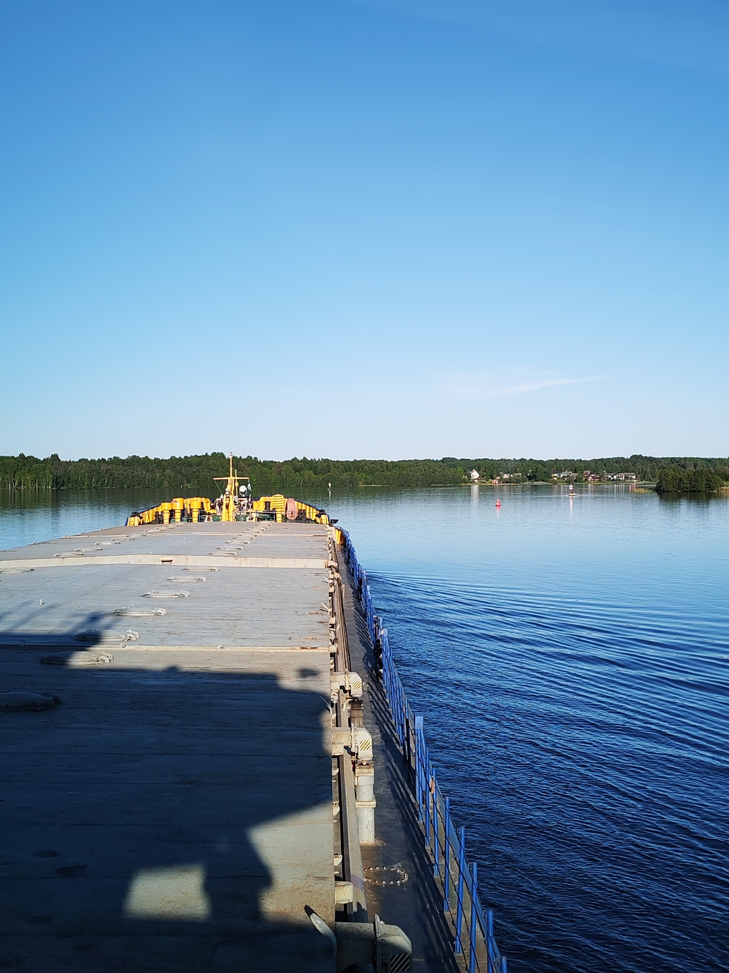 The image presents a daytime, panoramic view from the deck of a flat-topped vessel, likely a barge, traversing a large body of water in the vicinity of Mys, Russia. The vessel's grey deck, exhibiting several hatch covers and obscured shadows, occupies the lower left foreground. A blue railing lines the vessel's starboard side, adjacent to the water. The forward section of the vessel features a series of yellow cylindrical fenders and a central mast-like structure equipped with various attachments. The water is a deep blue, showing distinct ripples along the starboard side, suggesting forward motion. Two navigational aids are present in the water: a red conical buoy to the left and a white cylindrical buoy to the right, topped with a small flag or light. The background is dominated by a dense, dark green forest bordering the shoreline, behind which a cluster of residential structures with light facades and red roofs is discernible on the right. The sky overhead is uniformly clear and light blue, without any cloud formations. No specific human figures are distinctly visible.