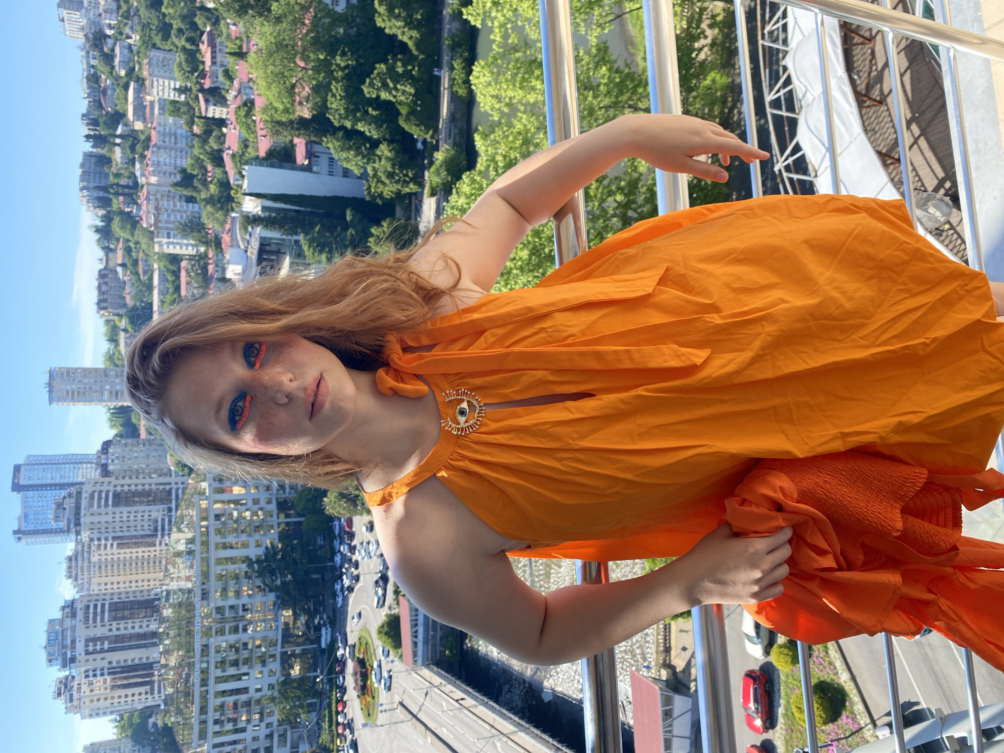 A young woman with red hair and bright blue and orange eye makeup stands on a balcony overlooking a city. She is wearing an orange halter dress with a gold evil eye brooch at the neckline. She is holding a crumpled orange fabric in her right hand. The background shows a densely built city with modern high-rise buildings and older houses, interspersed with lush green trees. In the distance, a body of water can be seen. The scene is set in Sochi, Russia, as indicated by the architecture and the typical urban landscape of a coastal city in the region. The sky is clear and blue.