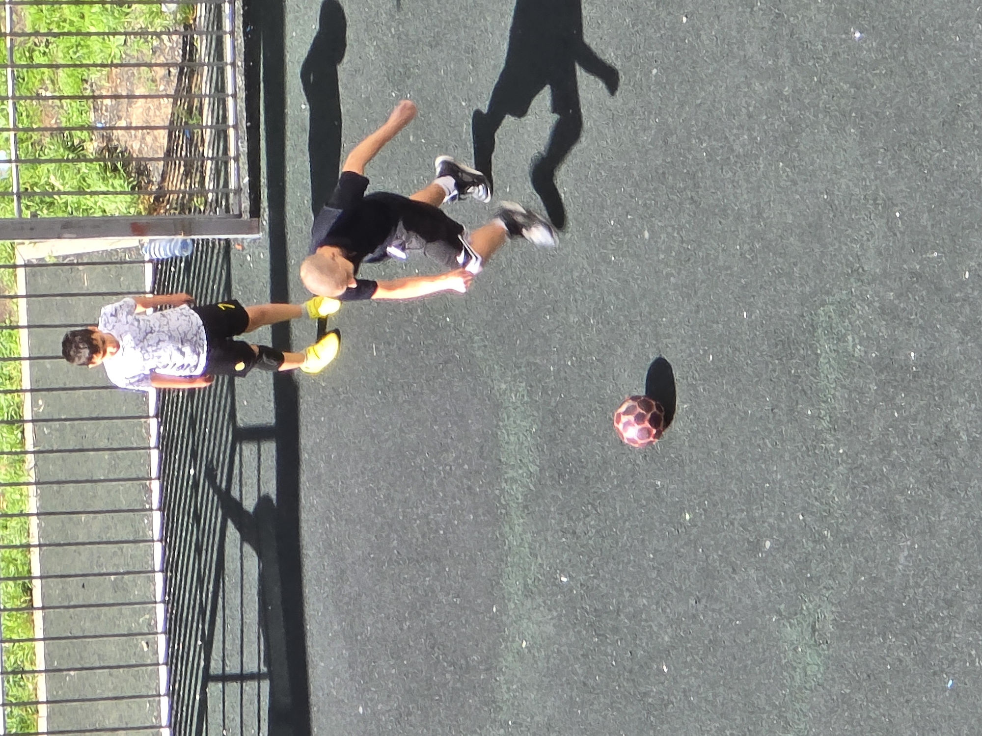 A high-angle shot shows two boys playing with a soccer ball on a gray-surfaced area. One boy wears a black shirt and shorts with white trim, along with white and black sneakers. The second boy has on a white and gray patterned shirt with black shorts and yellow shoes. The ball appears to be a small, red and black soccer ball. The boys are on an asphalt area, partially enclosed by a metal fence, next to a grassy area. A water bottle rests near the fence. Their shadows are cast on the ground. The location is Khimki, Russia.