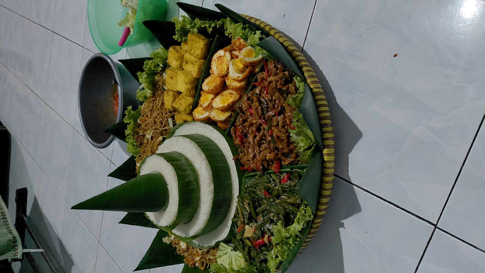 An overhead, high-angle shot shows a large, round, woven bamboo tray filled with Indonesian food, likely a Nasi Tumpeng. The tray is placed on a white tiled floor, with visible grout lines and a shadow cast by the food. The Nasi Tumpeng itself is a cone of white rice, layered and wrapped with green banana leaves, topped with a conical banana leaf decoration. Surrounding the rice are various side dishes, including stir-fried noodles, cubed fried tofu, sliced hard-boiled eggs, stir-fried long beans with chili, and stir-fried tempeh with chili. Green lettuce leaves are used as a garnish and to separate the different food items. In the background, a dark grey metal bowl and a light green plastic bowl are visible on the tiled floor. The lighting suggests an indoor setting, possibly a home in Pasirmuncang, Indonesia, given the presence of local cuisine.