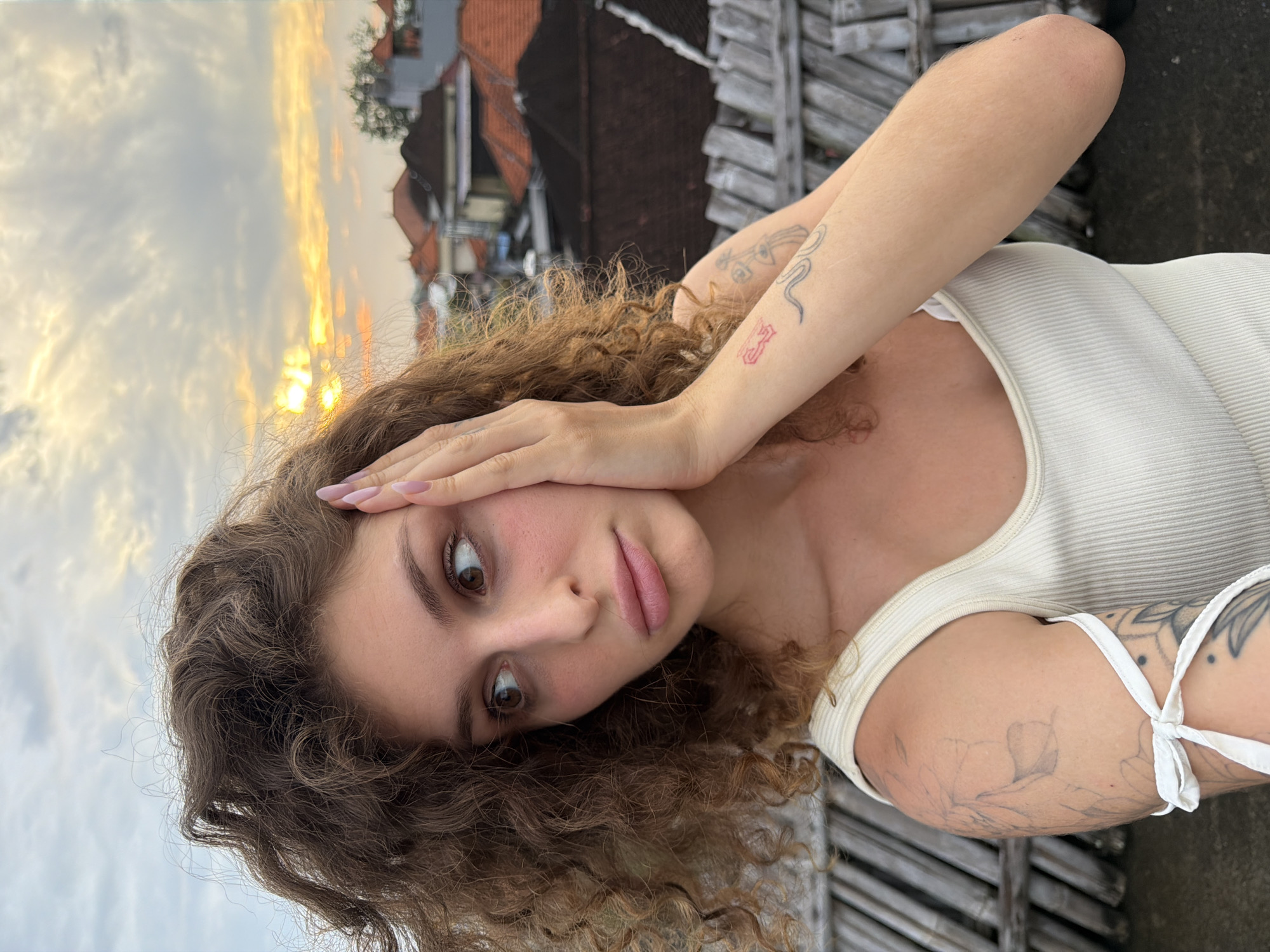 A young Caucasian woman with curly brown hair is pictured in a close-up shot, her right hand touching her face. She wears a white ribbed tank top and has tattoos on her arms. Her gaze is directed to her left, with a slightly surprised or concerned expression. In the background, a hazy sunset casts a warm glow over rooftops, suggesting an urban or suburban setting in North Kuta, Indonesia. The foreground features a weathered wooden structure, possibly a railing or fence.