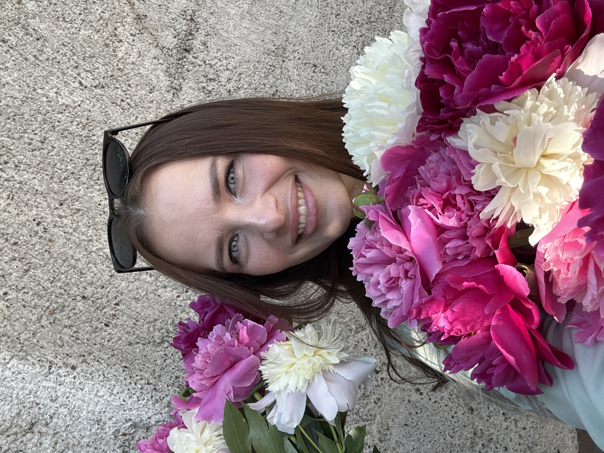 A woman with long brown hair, wearing sunglasses on top of her head, smiles at the camera. She is holding two large bouquets of peonies, one on her left and one in front of her. The flowers are a mix of pink, white, and deep fuchsia. The background is a rough, textured, gray wall. The scene appears to be outdoors in the city of Chaikovsky, Russia.