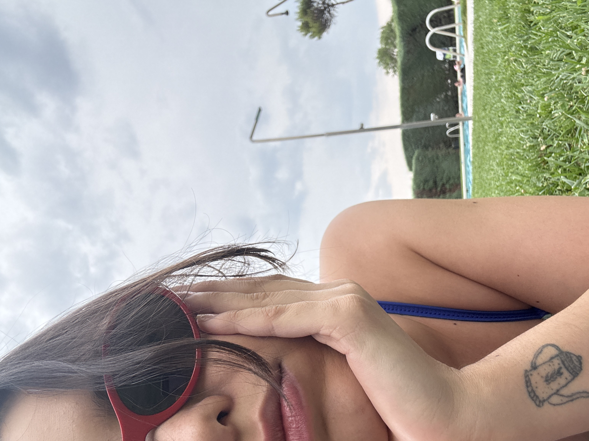 A person with dark hair and red sunglasses rests their head on their hand, close-up, on a grassy area. They are wearing a blue bikini top. A tattoo of a jug with a figure inside is visible on their wrist. In the background, a swimming pool with several people in it is visible, surrounded by trimmed hedges. Two lampposts stand near the pool, and a tree is visible in the distance. The sky is overcast. The location is in the city of El Escorial, Spain.