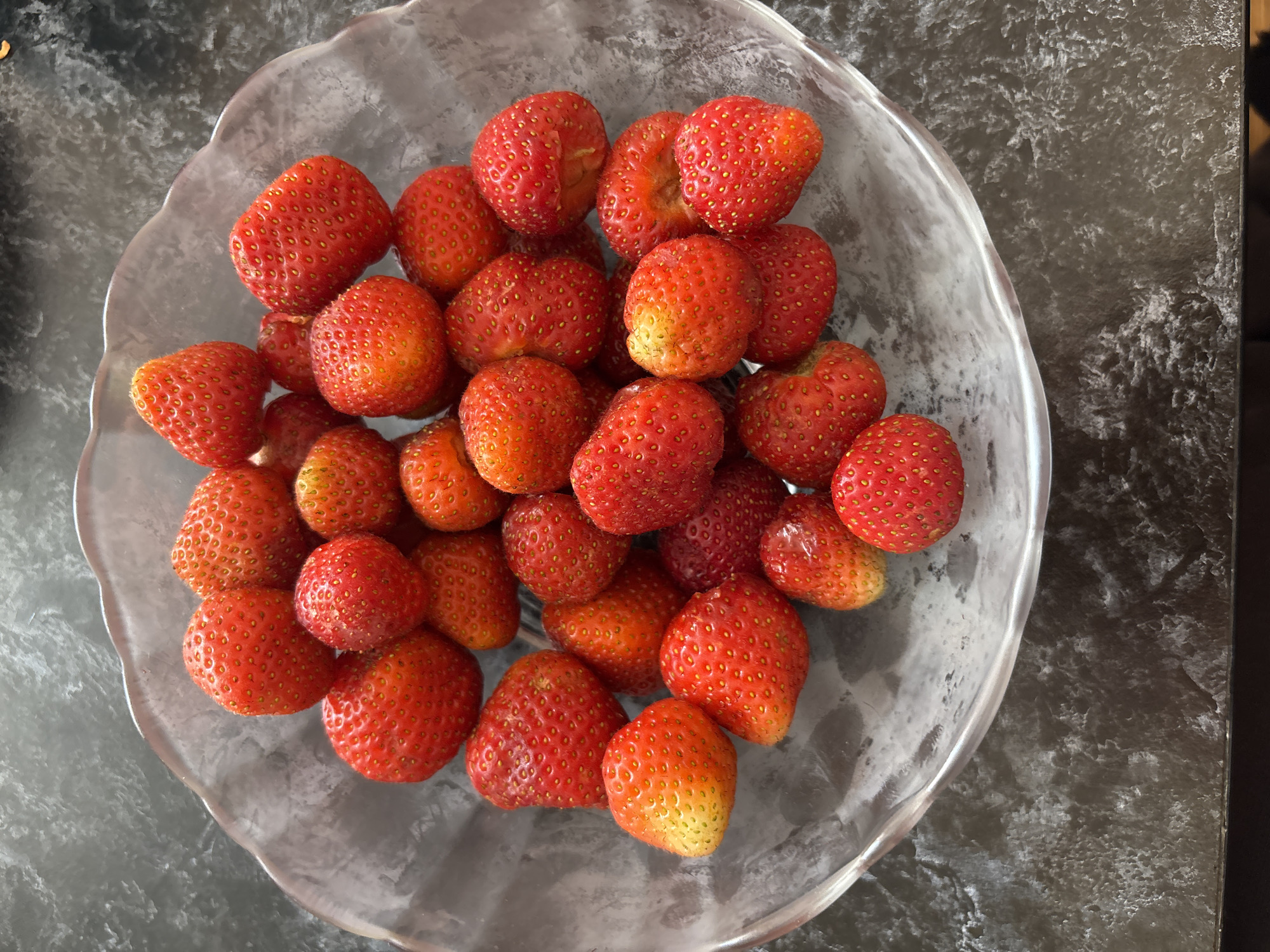 A clear, wavy-edged glass bowl filled with ripe strawberries sits on a dark, textured countertop. The strawberries are various shades of red and some have yellow-orange tinges, indicating ripeness. Their surfaces are covered in small, yellow seeds. The bowl is positioned slightly off-center in the frame, and the lighting casts subtle shadows, highlighting the contours of the fruit and the bowl's texture. The background is a dark, mottled surface, suggesting a table or counter. There are no people or animals present in the image, and no discernible city or location information is available.