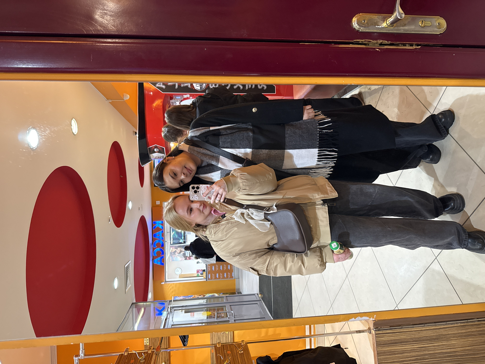 Two young women are depicted in a selfie taken in a mirror, likely within the lobby of the Kinovremya Cinema, located at Lenin Street, 39А, in Chaikovsky, Perm Krai, Russia.
The woman on the left, with blonde hair, wears a beige puffer jacket, dark grey wide-leg trousers, and black platform shoes. She holds a light-colored smartphone with a patterned case in her right hand, capturing the reflection, and a green-labeled bottle in her left hand. A dark brown crescent-shaped shoulder bag is slung across her body.
The woman on the right, with dark hair, wears a black coat, a black and white checkered scarf with fringe, dark trousers, and black platform shoes. She leans her head slightly towards the first woman, smiling.
The background, reflected in the mirror, reveals an interior space with bright orange walls and a white ceiling featuring several large, recessed red circular panels and recessed lighting. A prominent neon-blue sign displaying "KACCA" (Касса - Cashier) is visible, indicating a checkout area. A wall-mounted clock and a film poster are also visible near the cash register. To the left, a glass double door with a visible "ВЫХОД" (Exit) sign is reflected. A metal display rack is partially visible on the far left. A red structure with a black top, possibly a concession stand, is visible behind the woman on the right. The floor is covered with light-colored tiles and dark grout lines. A white dome-shaped CCTV camera is mounted on the orange wall. The right edge of the mirror is framed by a dark red-brown door with a brass handle and lock mechanism.