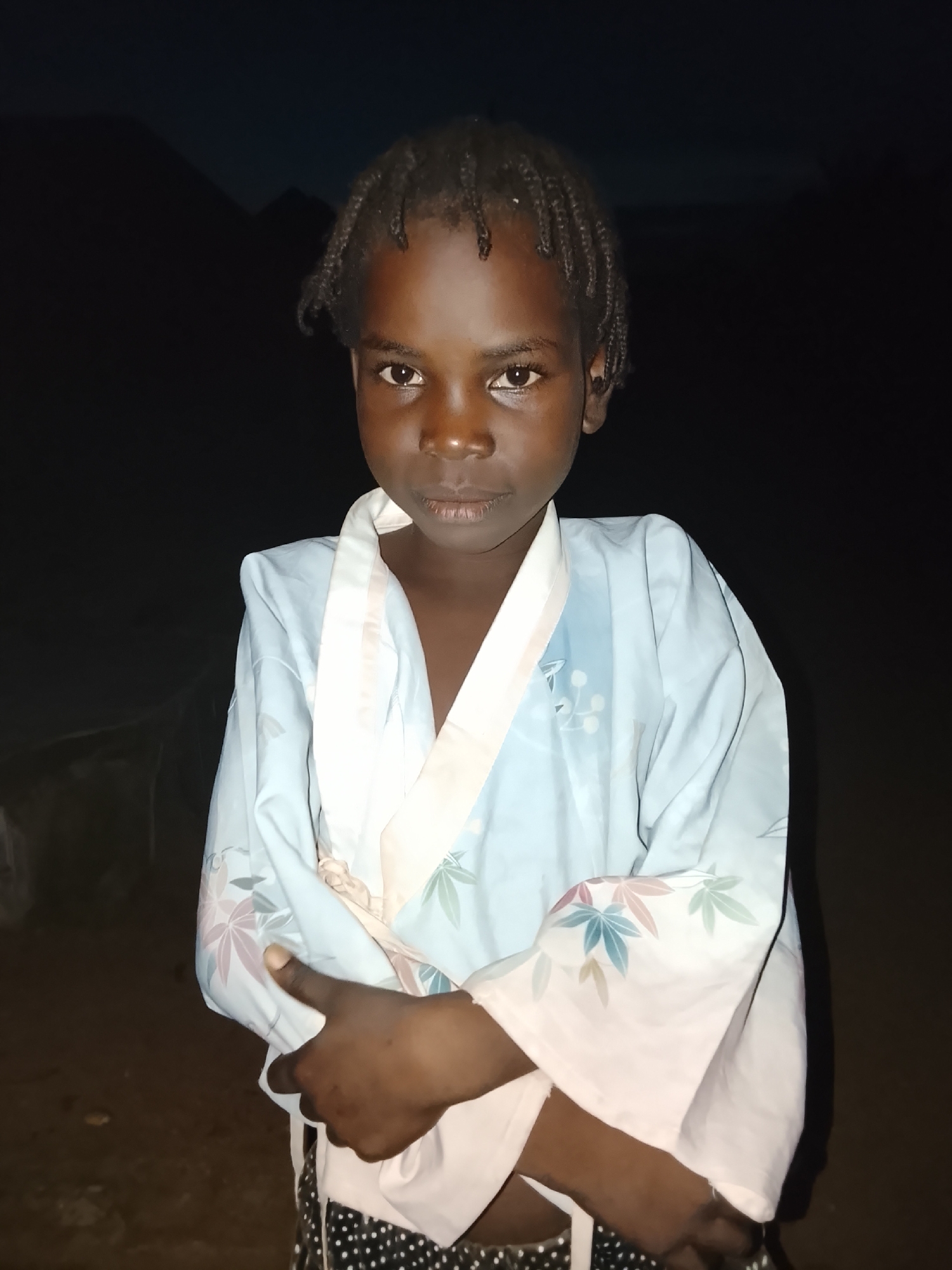A young Black person with braided hair is pictured standing in front of a dark, outdoor background that suggests it is nighttime. They are wearing a light blue robe with a white trim and a floral pattern featuring pink, blue, and green leaves. Their arms are crossed in front of their chest. The lower portion of their attire, visible beneath the robe, consists of black and white polka dot patterned shorts or pants. The scene appears to be in a rural or undeveloped area, as indicated by the dark, indistinct background that could be natural terrain. The image does not contain any elements that specifically indicate the location as Akwanga, Nigeria, beyond the prompt's assertion.