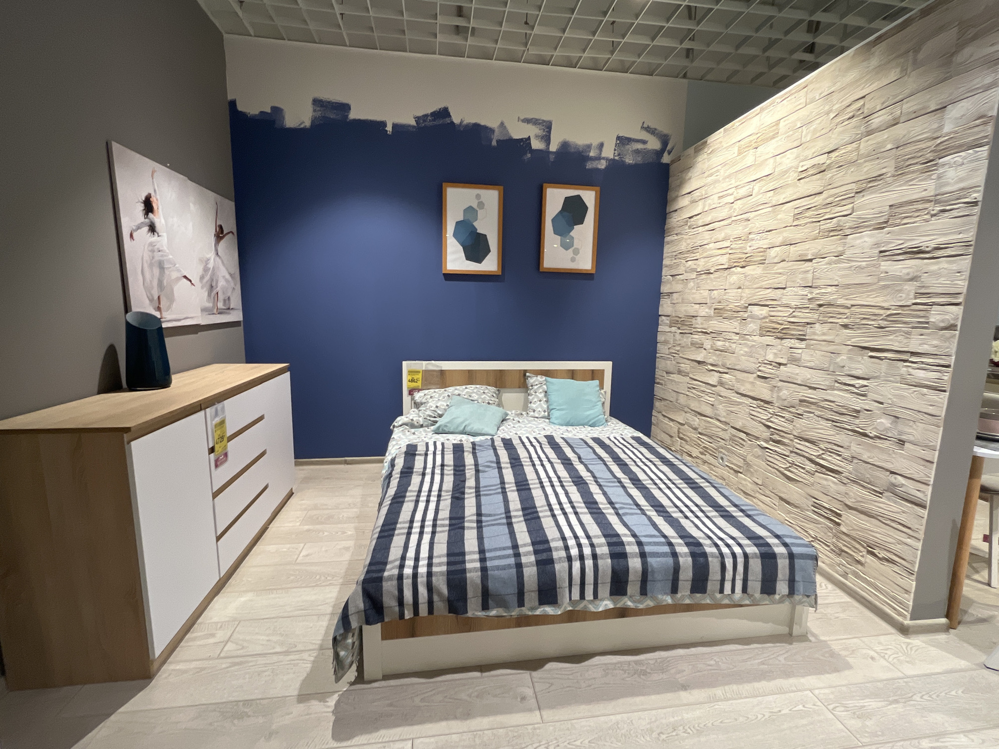 The image shows a bedroom display in a furniture store. The room is decorated with a dark blue accent wall, a light wood paneled wall, and a neutral ceiling. On the left side of the room, there is a light wood dresser with white drawers. Above the dresser, there is a large canvas painting of two ballerinas. A dark blue vase is placed on top of the dresser. In the center of the room, there is a bed with a wooden headboard and a white frame. The bed is covered with a blue and white plaid duvet and two light blue pillows. Two framed geometric art pieces hang on the blue wall above the bed. The floor is made of light-colored wood-look tiles. The overall impression is of a modern and stylish bedroom setup. The location is a furniture store in Kharkiv, Ukraine. There are no people visible in the image.