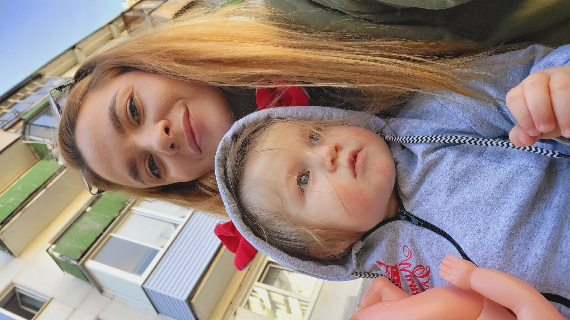 A woman and a child are posing for a selfie against the backdrop of an apartment building. The woman has long blonde hair and is smiling. The child, wearing a gray hooded sweatshirt with a red bow attached to the hood, is looking upwards. A partial view of a pink-colored toy is visible. The building has balconies with green and blue paneling. The location is identified as the city of Chaikovsky, Russia.