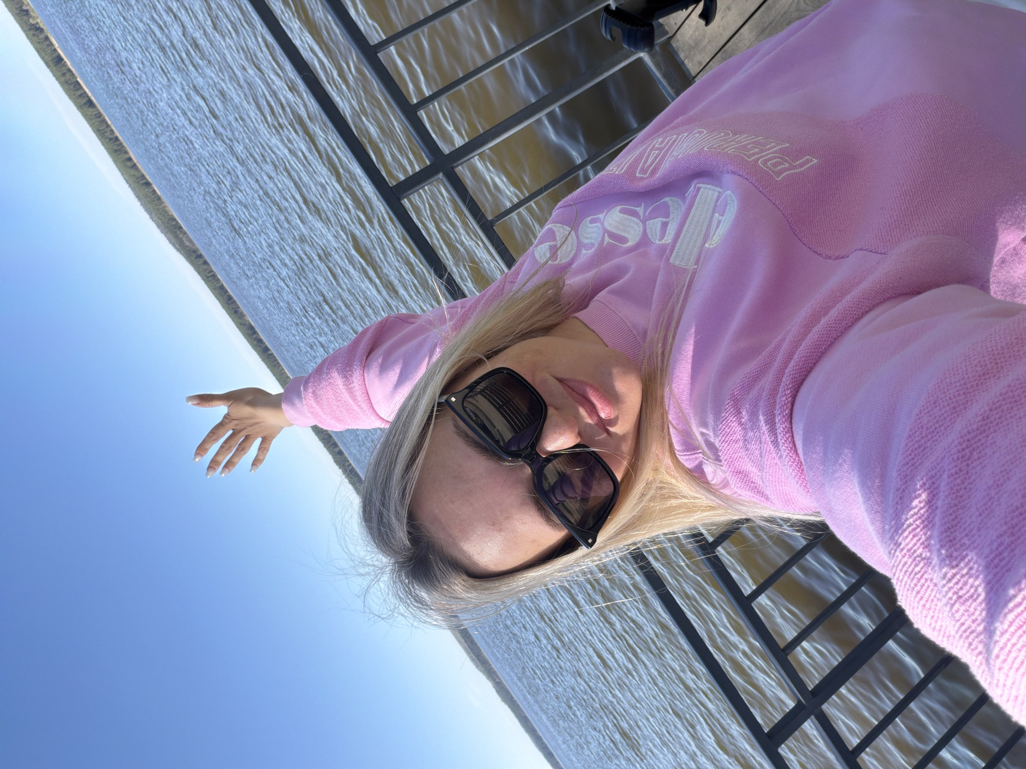 A woman with blonde hair is taking a selfie outdoors. She is wearing sunglasses and a pink sweatshirt with white lettering. She is standing on a metal structure, possibly a dock or a bridge, with water visible in the background. The water appears to be part of a large body of water like a lake or reservoir, stretching towards a distant shoreline with trees. The sky is a clear blue. The woman has one arm extended towards the sky. It is plausible that the photo was taken in or near Votkinsk, Russia, given the water and shoreline features resemble the Votkinsk Reservoir.