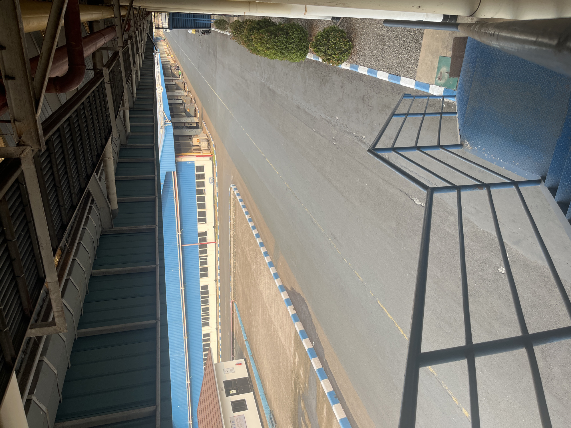 The image depicts an outdoor industrial area in the city of Pelang, Indonesia. In the foreground, a blue metal staircase with railings leads down. Beyond the staircase, an asphalt road stretches into the distance, bordered by a white and blue curb. To the left of the road, there is a small building with a brown roof and a tan-colored yard. Further down the road, a long, blue-colored building with numerous windows is visible. The area is lined with several evenly spaced green bushes. Overhead, a covered walkway with exposed metal beams and pipes is present, suggesting an elevated pedestrian or service pathway. The lighting indicates it is daytime.