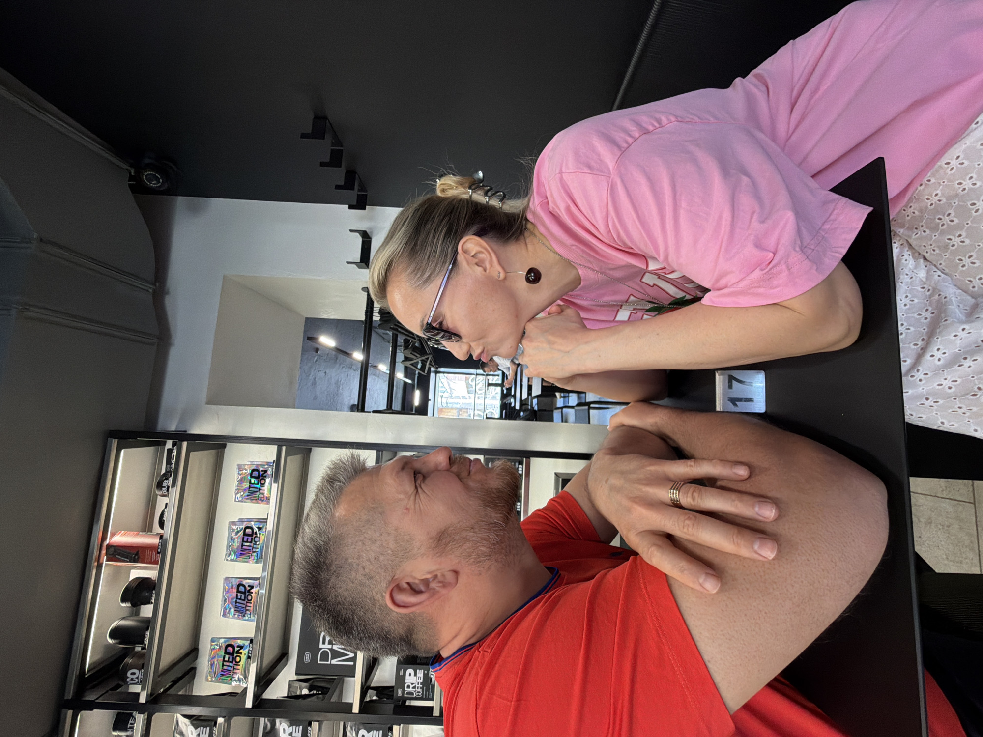 Here is a description of the image:
The image captures a close-up of two individuals, seemingly a man and a woman, leaning towards each other over a dark table. The woman is positioned on the right side of the frame, wearing a pink shirt and glasses. The man is on the left, sporting a red shirt and a wedding ring on his left hand.
They are in a space with shelves displaying various items, possibly merchandise. The wall behind the shelves is white, contrasting with the dark ceiling above. A sticker reads "DRIP COFFEE". A sign on the table says "17". The location is a store called "Dream Makers" in Izhevsk, Russia.