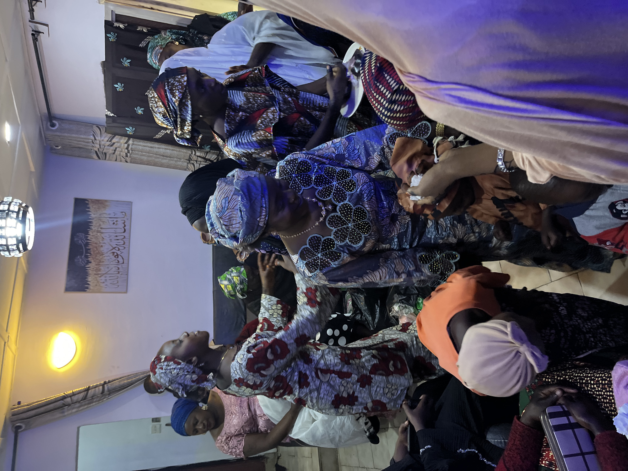 The image depicts an indoor gathering, likely a social event or celebration, within a residential setting in Jos, Nigeria. A group of women, predominantly dressed in colorful traditional African attire, are present. One woman in the foreground, wearing a patterned blue and white dress and headscarf, is looking downwards and appears to be handling something with her hands. Another woman beside her, in a pink lace top and blue headscarf, is looking to the side. Further back, other women are visible, some wearing hijabs. In the background, there are curtains and a framed artwork on the wall. The lighting is warm, with a prominent overhead light fixture and some purple ambient lighting casting shadows. The overall atmosphere appears festive or commemorative, given the attire and gathering of people.