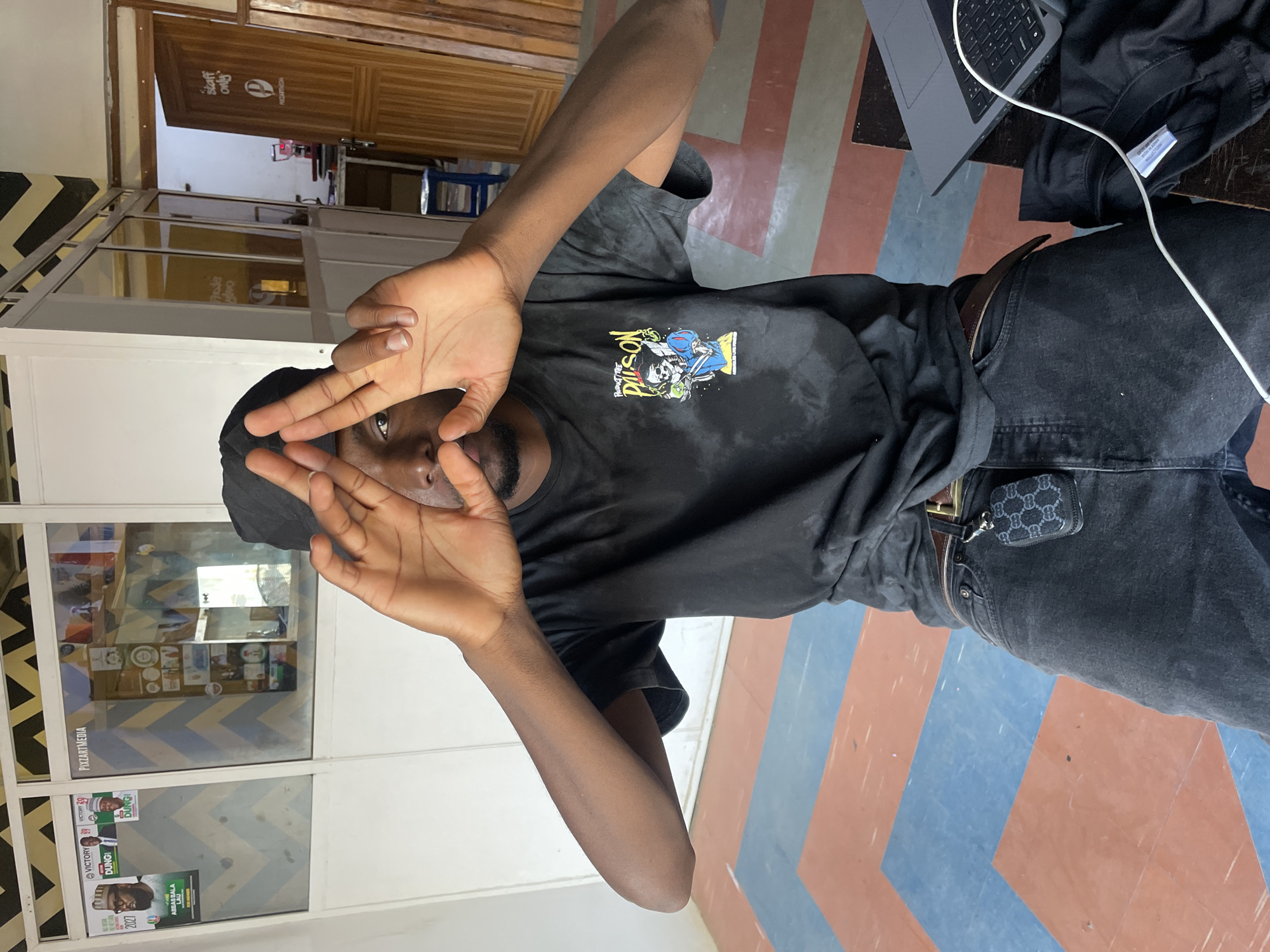 An indoor, vertical shot features a young man in Jos, Nigeria, posing directly for the camera. He is centered in the frame, holding his hands up to partially obscure and frame his face, looking through his fingers. He wears a black t-shirt with a colorful graphic design on the chest, depicting a skeleton and another figure with the text "PRODUCT THE PISON." He also wears dark jeans with a brown belt and has a small, black patterned pouch (resembling a designer AirPods case) clipped to a belt loop. A black cap or head covering is visible on his head.
The setting appears to be a commercial or office space. To the man's left, a glass partition displays various items and text, including "PIXZARTMEDIA" and several political-style posters featuring faces and text such as "VICTORY 39," "DUNGI," "2027," and "AMBASS BALA LAU." Further left, a wooden door with "staff only" written on it, along with a logo and the text "MEZARTHIGIA," suggests a restricted area. To his right, a grey laptop with a white charging cable rests on a dark surface. The floor beneath him has a distinctive geometric pattern of reddish-brown and blue shapes arranged in chevrons. The lighting is consistent with an indoor environment, likely artificial. The man's expression is neutral and direct.