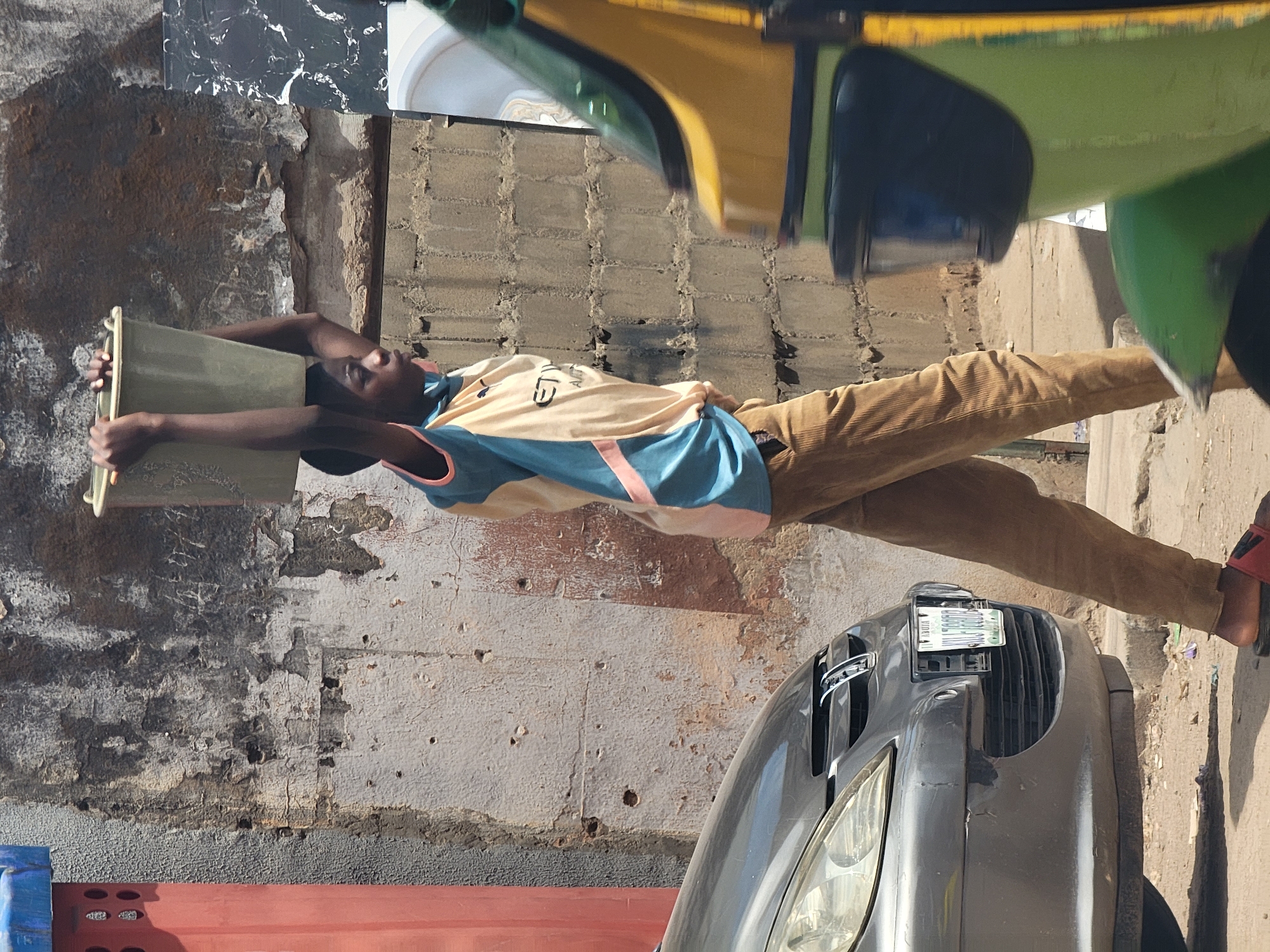 A young man, likely an adolescent, is carrying a rectangular, pale green container overhead. He is standing on a street in Maiburiji, Nigeria. He wears a yellow and blue sleeveless shirt and brown corduroy pants. The street has a gray car parked along the curb, as well as a red container. There is a green auto-rickshaw parked to the right. The background consists of a weathered concrete wall.
