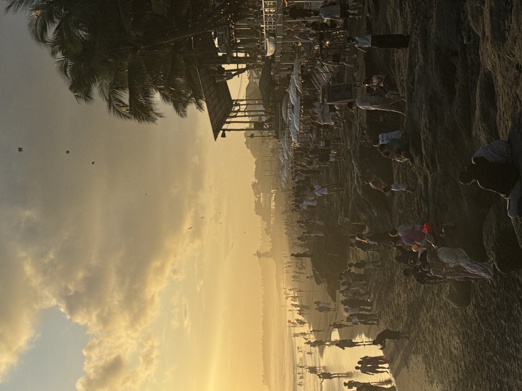 The image depicts a crowded beach scene in North Kuta, Indonesia at sunset. The sky is filled with golden light and clouds. Palm trees are visible along the shoreline and a building is present in the background. Many people are gathered on the beach, some are walking along the water's edge, others are standing or sitting. The beach itself is sandy and wet.