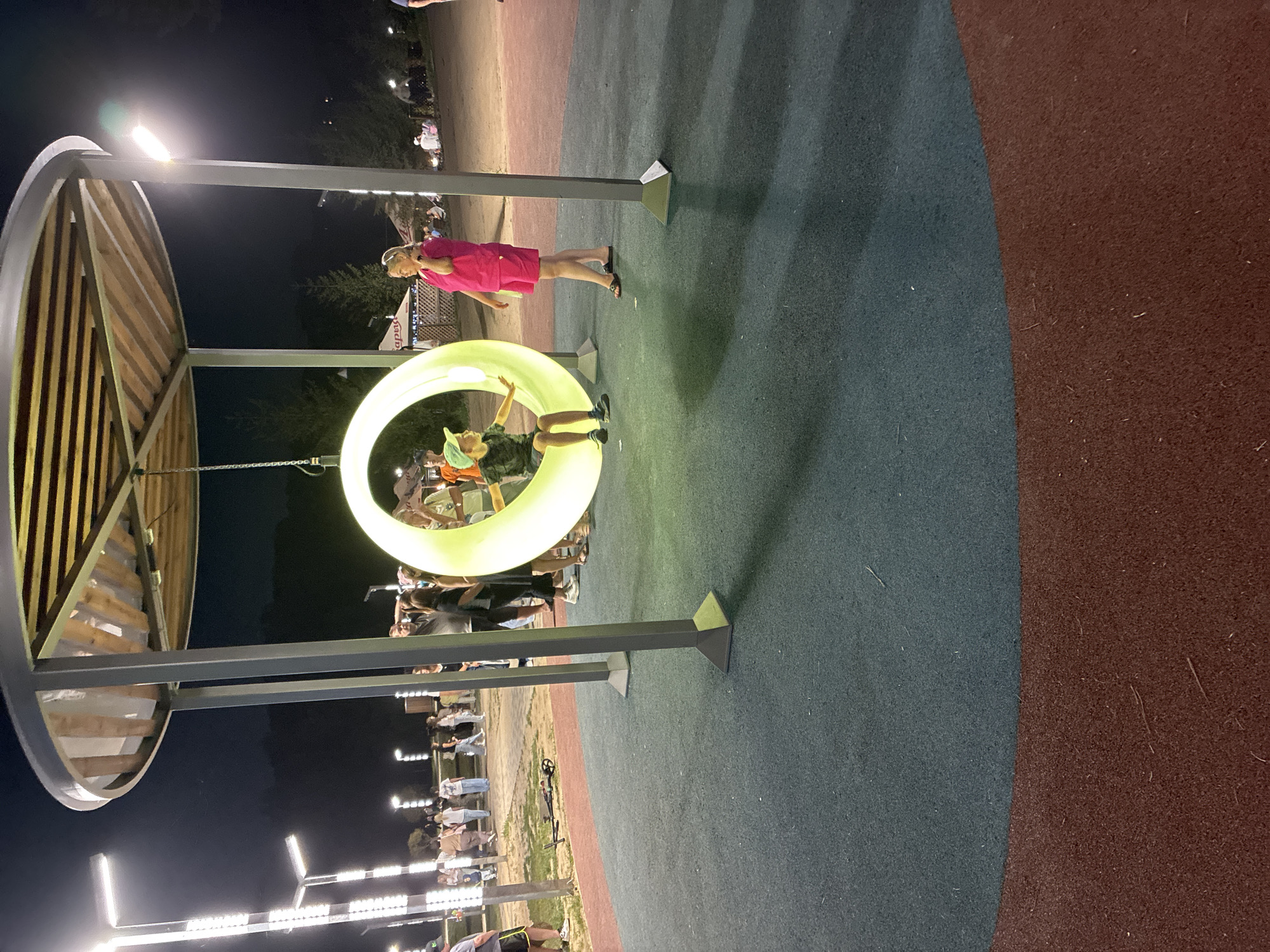 The image captures a nighttime scene at a park or playground in Mahilyow, Belarus. A large, illuminated circular swing, glowing with a neon green light, is the central focus. A young boy wearing a green cap and dark clothing is seated on the swing, appearing to be playing or being pushed. To the right of the swing, a woman in a bright pink outfit stands watching. In the background, a crowd of people are visible, some walking and others gathered, suggesting a public event or a lively evening atmosphere. The park features modern lampposts casting bright light, and the ground is covered with a soft, rubberized surface in contrasting blue and red sections. Trees and some structures, possibly stalls or tents, are discernible in the distance, contributing to the outdoor, recreational ambiance.