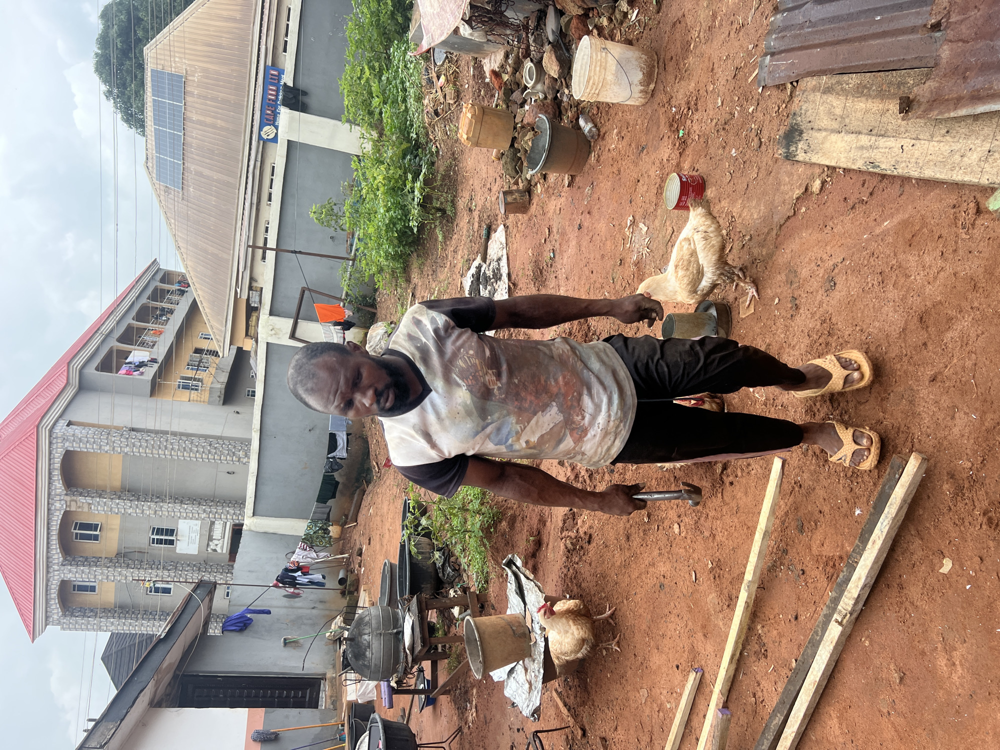 In Nkpor, Nigeria, a man stands in a dusty courtyard. He is wearing a t-shirt and dark pants with yellow flip-flops. In his right hand, he holds a hammer. A light-colored chicken walks on the red dirt ground to his right, and another chicken is visible to his left, near a pile of cloths laid out on the ground. In the background, there are several large, dark containers, some wooden structures, and a two-story building with a red roof. The sky is overcast.