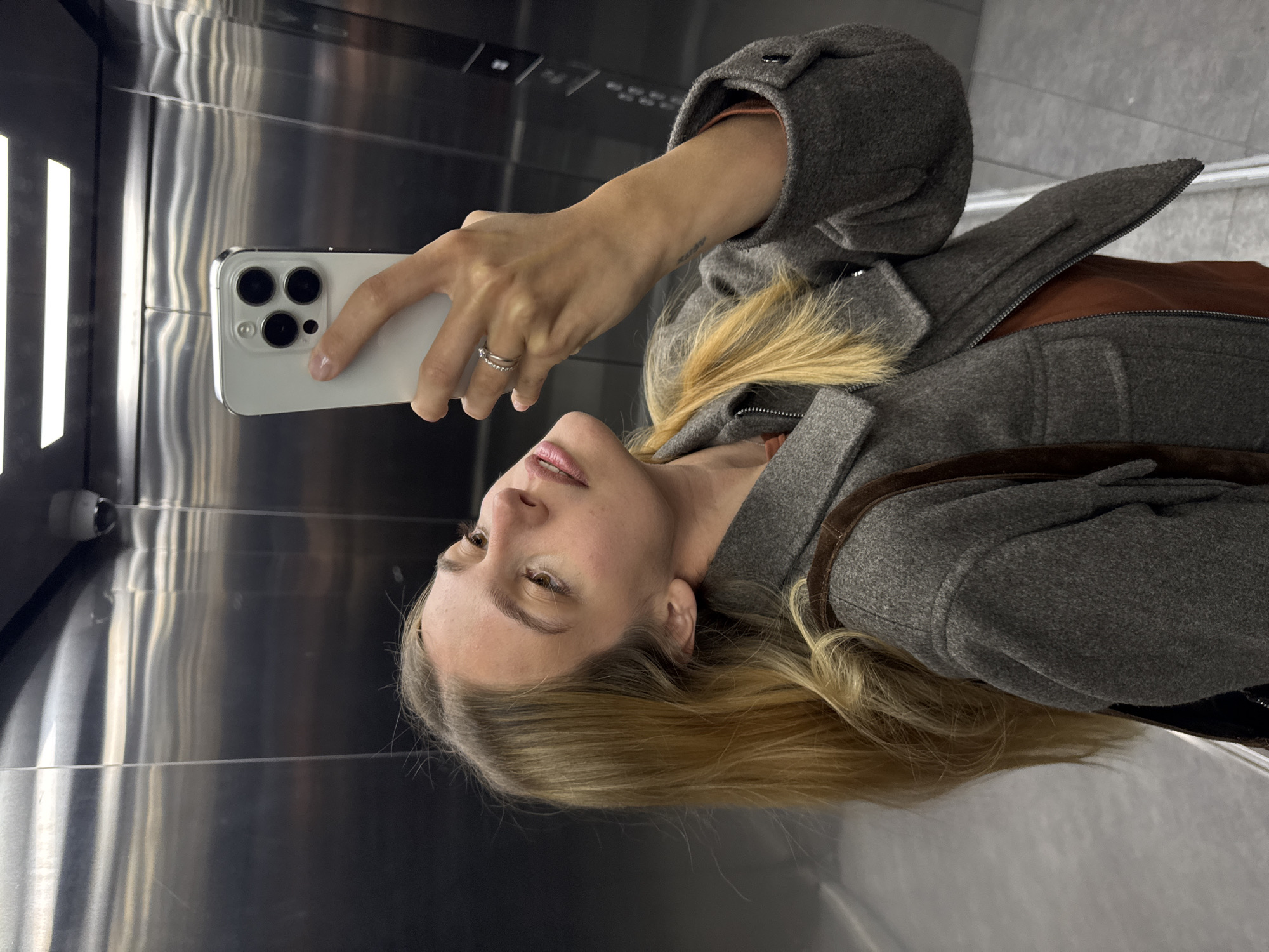 A woman is taking a selfie in an elevator. She is holding a white smartphone with three camera lenses. The elevator has stainless steel walls and a surveillance camera is visible in the upper left corner. The woman is wearing a gray wool coat with brown leather trim and a silver ring on her left hand. Her blonde hair is loose and wavy. She appears to be looking at the screen of her phone. The location is specified as Unknown, Unknown.