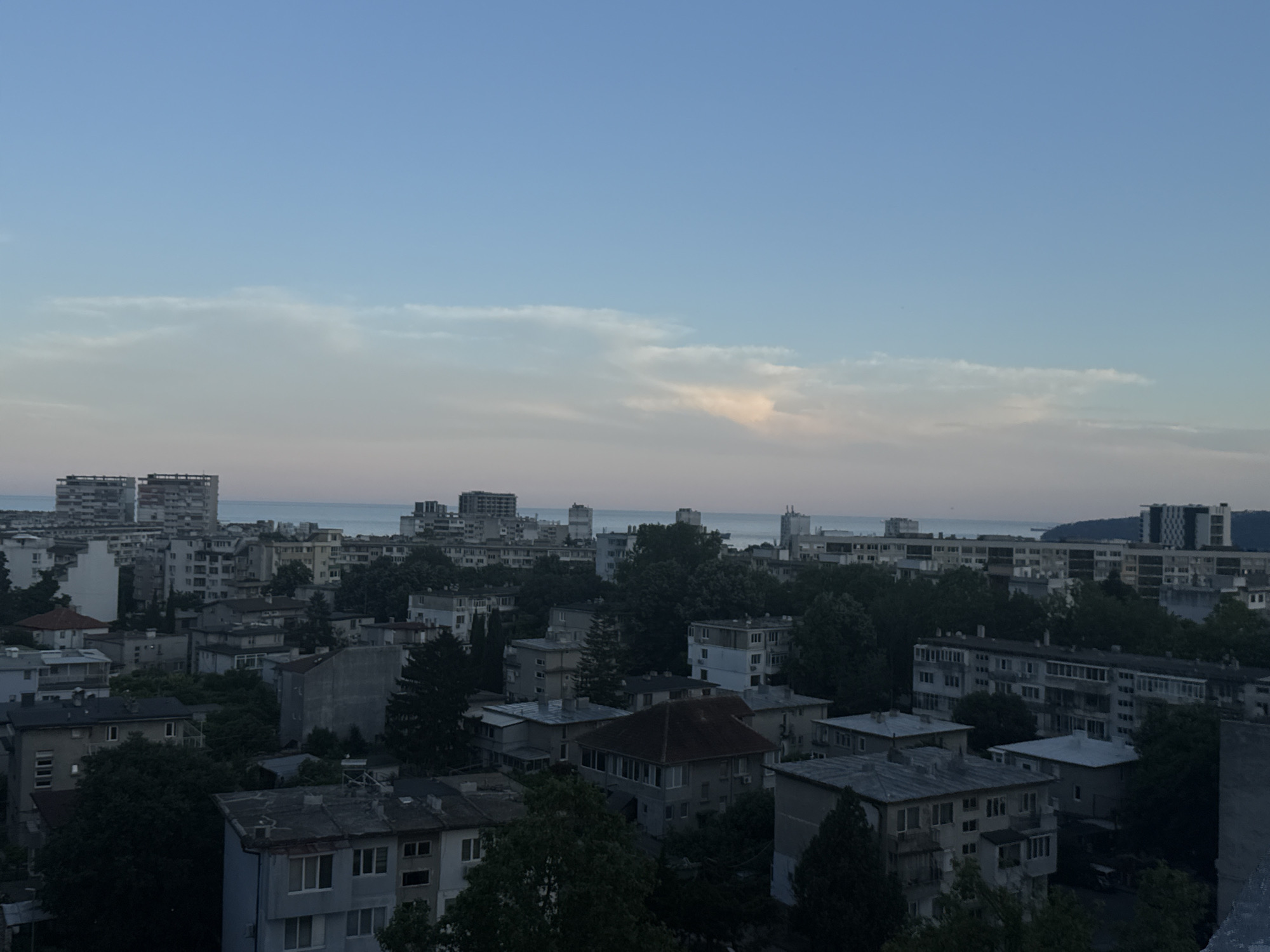 This is a wide-angle, high-angle shot of a city skyline at dusk. The sky is a pale blue with streaks of pink and orange clouds towards the horizon. Below the sky, the city is densely packed with buildings of varying heights and architectural styles. Many of the buildings appear to be apartment blocks, with repetitive window patterns. Some of the buildings are darker than others, suggesting they are in shadow, while others are lighter, possibly reflecting the remaining daylight. There are also many trees scattered throughout the city, some appearing quite large and leafy, contributing to a green canopy in places. In the distance, a body of water, likely the Black Sea, can be seen, with a few more distant buildings on the far shore. The overall impression is one of an urban environment at the end of the day. Based on the architectural style of the buildings, particularly the large apartment blocks, and the coastal setting, this scene is likely located in Varna, Bulgaria. The image captures a panoramic view of the city from a high vantage point.