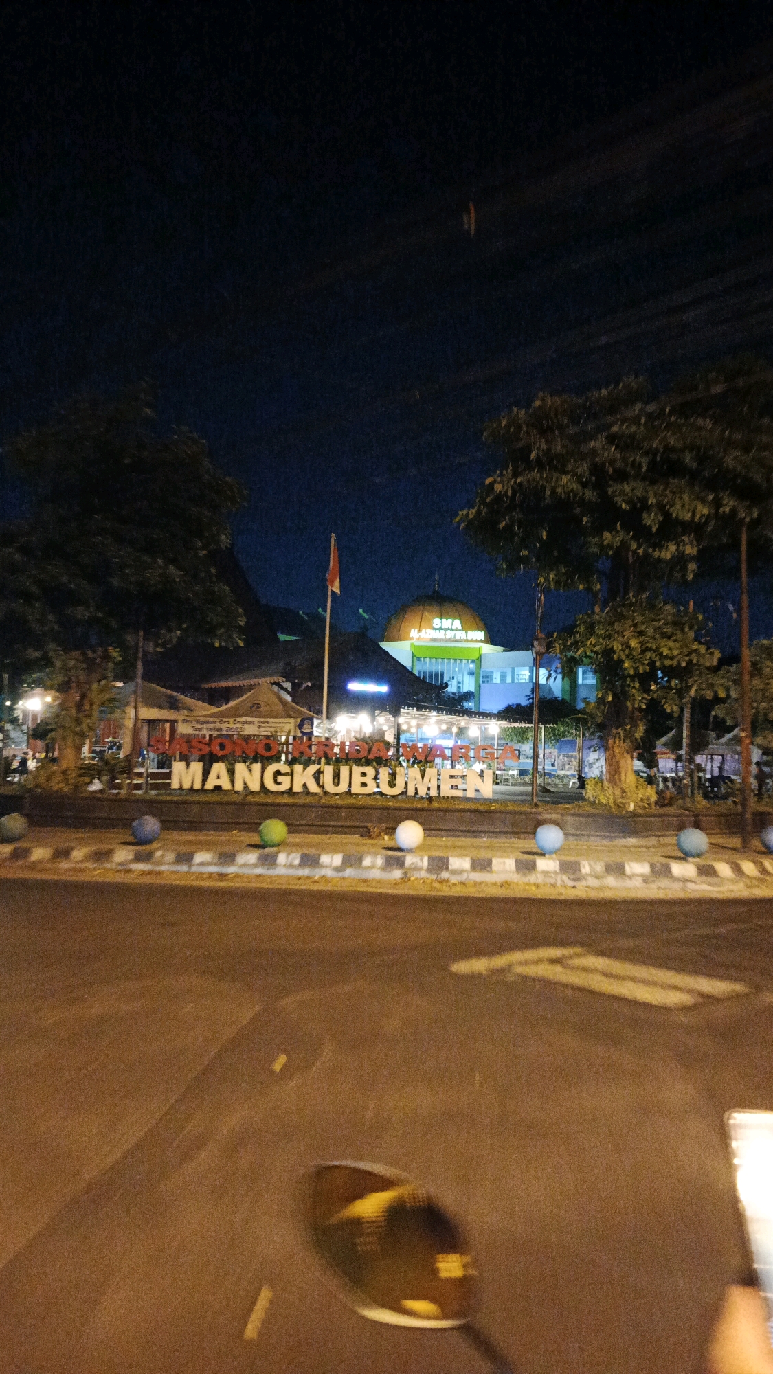 The image captures a night scene in Surakarta, Indonesia, featuring a prominent sign that reads "MANGKUBUMEN" in large white letters, with smaller text above that appears to be "SONO KRIDA WARGA". Behind the sign, a building with a green dome and "SMA AL-AZHAR SYIFA BDI" written on it is visible, illuminated by lights. A red and white flag flies on a pole in front of the building. The foreground shows a street with decorative spherical objects placed along the curb. Trees are present on either side of the scene, and the road surface is visible, reflecting some of the ambient light. The overall impression is of a public space or landmark at night.
