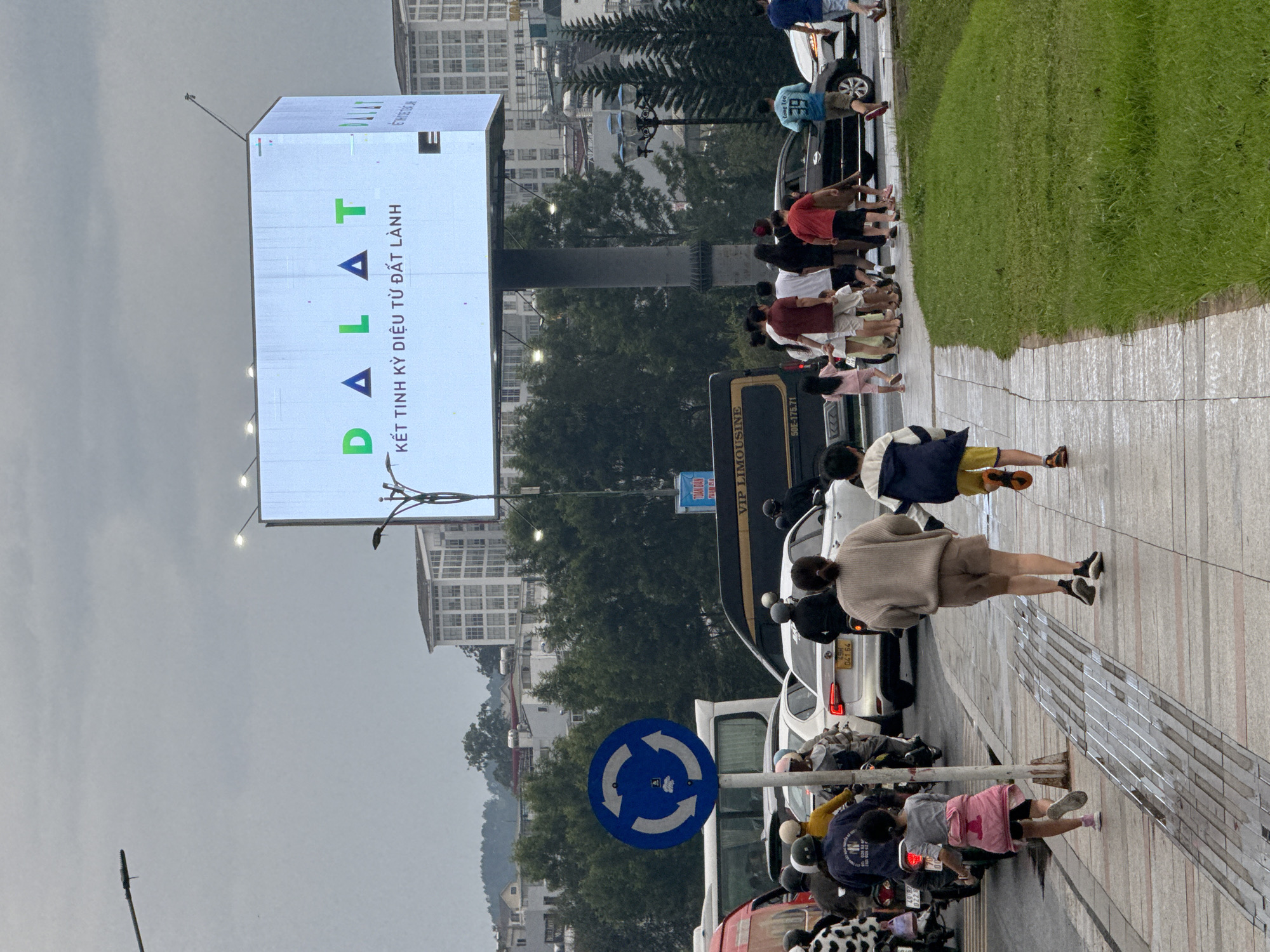 The image captures a street scene in Da Lat, Vietnam, under an overcast sky. A large digital billboard displaying "DALAT" in colorful letters and Vietnamese text "KẾT TINH KỶ DIỆU TỪ ĐẤT LÀNH" is prominent in the upper portion of the frame. Below the billboard, a busy street is filled with traffic, including motorcycles, cars, and a VIP Limousine bus. Pedestrians are visible on the sidewalk, with several children running. A blue circular traffic sign with white arrows indicating a roundabout is also present. The environment includes trees and buildings in the background, suggesting an urban setting. The ground appears wet, indicating recent rain.