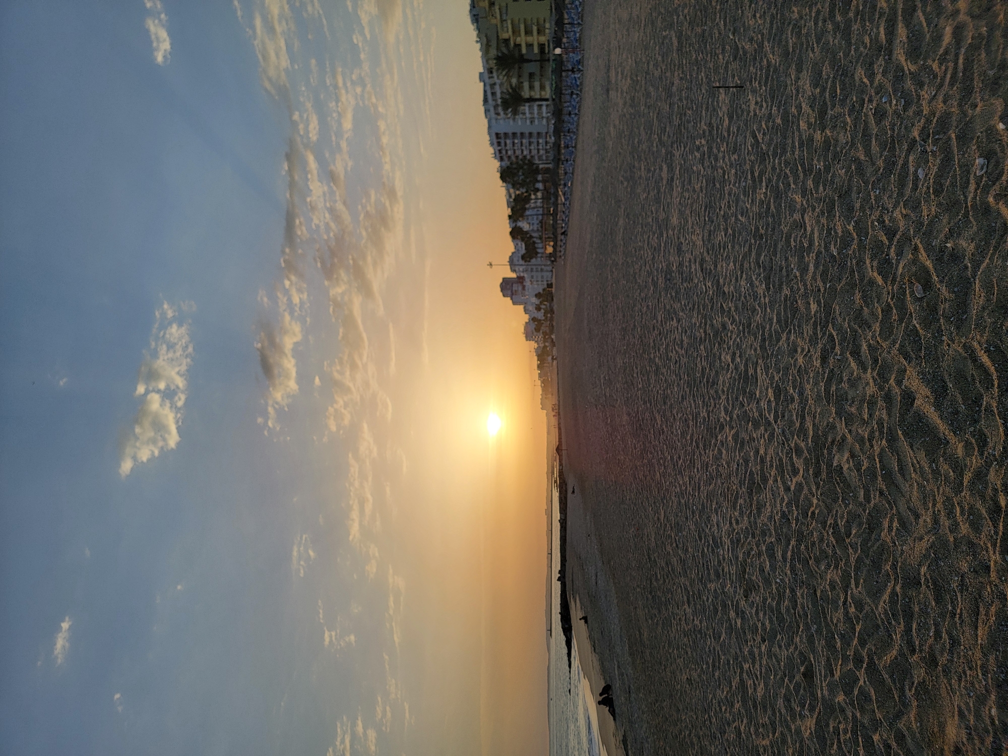 This is a vertical, full-frame photograph of the beach in Quarteira, Portugal, taken at sunset. The foreground is dominated by a wide expanse of sand with rippled textures from the receding tide. In the middle ground, the ocean meets the shore, with a rocky breakwater extending into the water on the left side. A few distant figures are visible sitting on the sand near the water's edge.
The background features a line of white buildings with multiple stories and balconies, typical of a coastal resort town, interspersed with palm trees. Beyond the buildings, the sun is setting on the horizon, casting a warm, orange glow across the sky and the scene. Scattered clouds, tinged with the sunset colors, dot the sky. The overall atmosphere is serene and peaceful, capturing the end of the day on the Algarve coast.