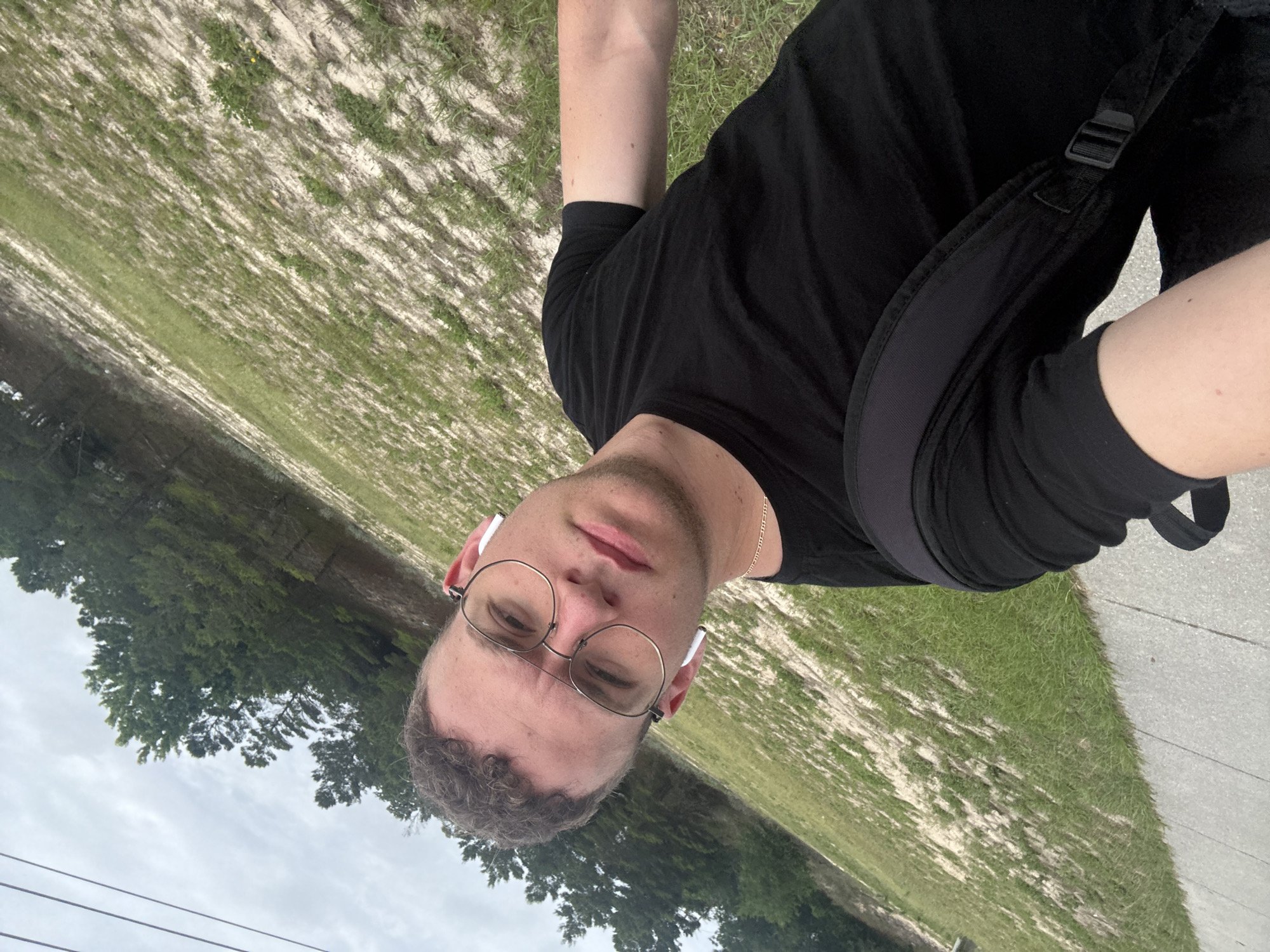 A selfie shows a Caucasian male wearing a black T-shirt, round metal-framed glasses, and white wireless earbuds. He is wearing a black backpack. The background shows sparse grassy areas adjacent to a paved path. Tall trees are present in the distance. The location is possibly Panama City Beach, United States of America.