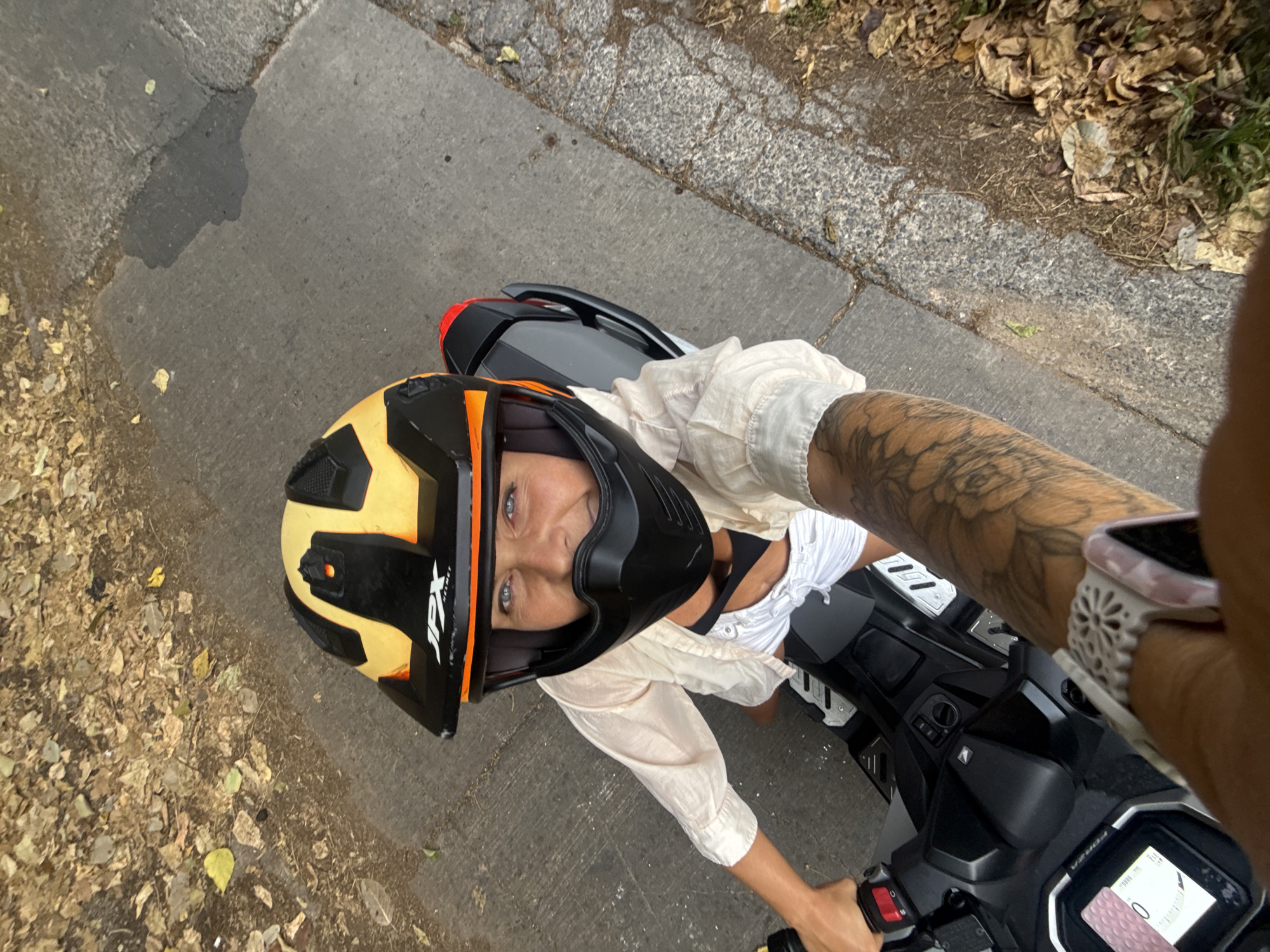 A woman wearing a yellow and black motorcycle helmet and a light-colored shirt over a black top and white shorts is seated on a black scooter. She is looking at the camera with a smile. Her left arm, which is tattooed, is extended towards the camera. The scooter is parked on a paved surface next to a dirt and leaf-covered embankment. The scene appears to be in an outdoor setting, possibly a roadside.
