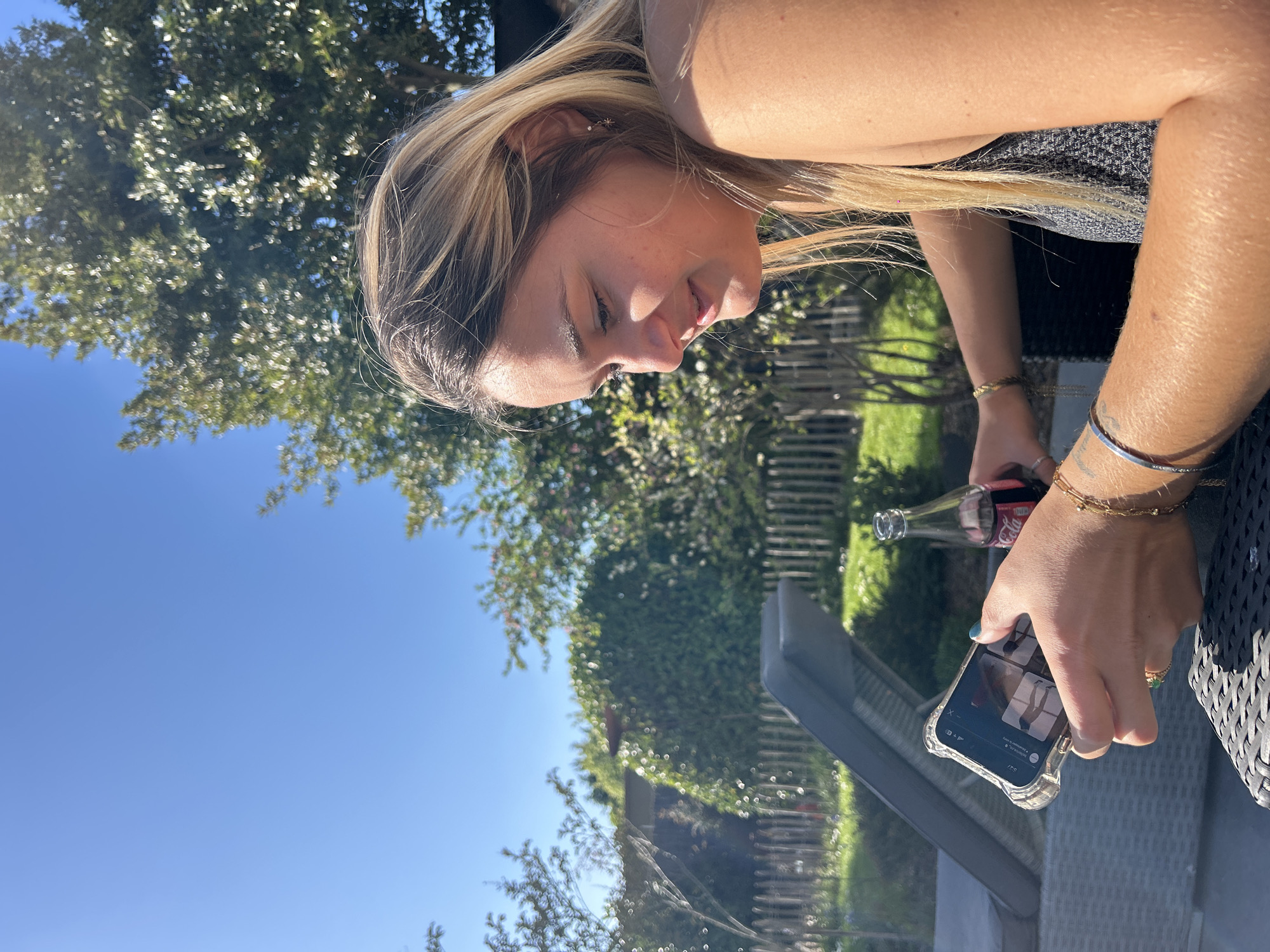 This outdoor image, taken in Toufflers, France, depicts a young blonde woman engaged with her smartphone on a bright, sunny day. The woman is shown in profile, looking down at her phone with a soft, serene expression and a slight smile. Her hair is illuminated by strong sunlight, which also highlights her skin.
She is seated or leaning at what appears to be outdoor furniture, visible on the right. In her right hand, she holds a glass Coca-Cola bottle, its label partially visible. Her left hand holds a smartphone, encased in clear plastic, which displays a social media or content browsing interface. The screen shows an "X" button at the top left, text that reads "recommended to you," and several small rectangular image previews, including what appears to be fashion or lifestyle content (e.g., legs, a standing person). The word "IT'S" is clearly visible at the bottom of the screen.
The woman wears several accessories: multiple bracelets on both wrists (gold chains, a silver band, beaded styles), rings on her fingers (one featuring a green stone), and a small star-shaped earring. Her fingernails are painted a teal or blue color.
The background features lush green trees and foliage, with a clear blue sky indicating fine weather, likely mid-day to late afternoon due to the bright sunlight. A wooden fence is partially visible behind the greenery. The scene conveys a relaxed and casual outdoor setting.