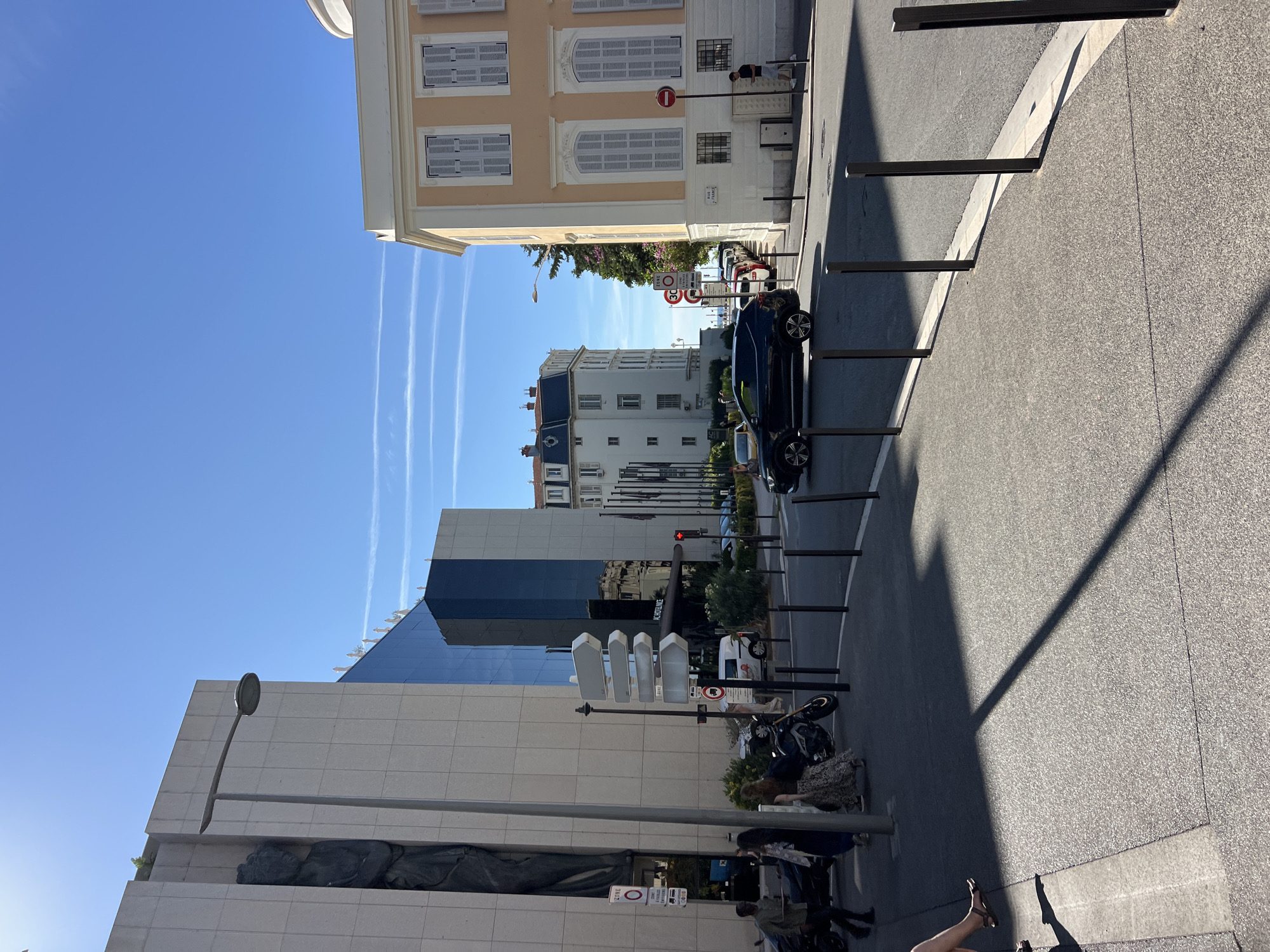 This is a street scene in Nice, France, on a sunny day with a clear blue sky and contrails from airplanes. The foreground shows a sidewalk with a gravel texture and a distinct shadow cast by an unseen object. A person's legs and feet, wearing sandals, are visible on the left edge of the frame, sitting on the sidewalk.
Moving towards the street, there are several black bollards lining the edge of the sidewalk. A dark-colored SUV is driving on the asphalt road, casting its own shadow. A motorcycle is parked on the sidewalk. A street lamp is positioned on the left side of the frame.
In the background, there are several buildings. On the left is a modern, light-colored building with large vertical panels and a dark sculpture integrated into its facade. Adjacent to it is a building with a dark, reflective glass exterior. Further down the street, a white building with a dark roof is visible, and on the right, a pastel yellow building with ornate window shutters and decorative trim stands prominently.
Traffic signs are visible, including a "ZONE 30" speed limit sign and other directional and regulatory signs. People are walking on the sidewalk, some near the motorcycle and others further down. A sign for "AC HOTEL NICE" is partially visible. The overall environment suggests a well-developed urban area with commercial and residential buildings.