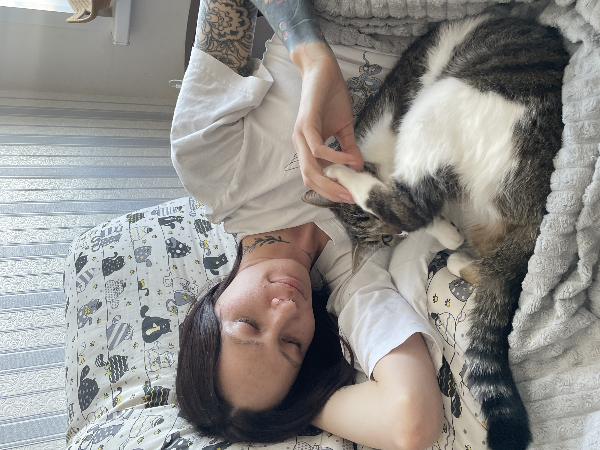 A female individual is depicted resting in a domestic setting in Izhevsk, Russia. She lies on her left side with her eyes closed, possessing dark hair, two rings piercing her right nostril, and visible tattoos on her neck (a leafy branch) and both arms (complex floral and ornamental designs, with the left arm featuring darker, denser ink). She wears a white short-sleeved t-shirt. Adjacent to her, a domestic cat, predominantly white with brown and grey tabby markings and a distinctly striped tail, rests on a light grey textured blanket. The woman's right hand is observed gently interacting with the cat's front paw. They are positioned on a bed adorned with a pillowcase featuring a pattern of stylized cats. The background wall is covered in wallpaper characterized by alternating vertical grey-blue stripes and white sections with a subtle patterned texture. A window sill, supporting a small woven object, is visible in the upper right quadrant.