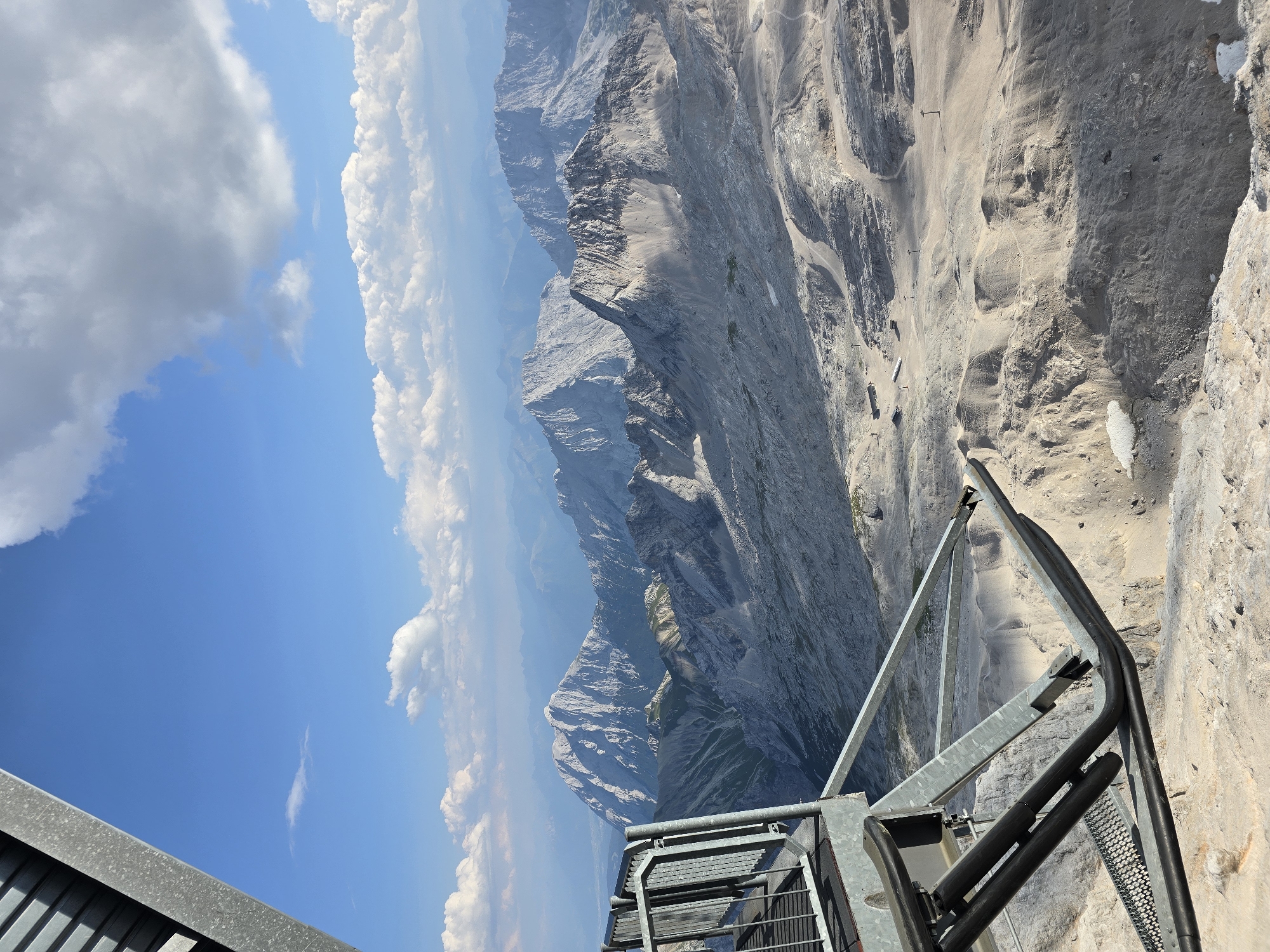 This image captures a panoramic view of the Zugspitze mountain range, the highest peak in Germany, located near Garmisch-Partenkirchen. The foreground features a metal structure, likely part of a viewing platform or cable car station, with a grey railing and diagonal metal supports. This structure juts out over a vast, rugged mountain landscape. The terrain is characterized by rocky slopes, scree, and some patches of snow. In the mid-ground, several mountain peaks are visible, with jagged ridges and shadowed valleys. The furthest mountains appear hazy and are layered in shades of blue, indicating distance. The sky above is a vibrant blue with scattered white and grey clouds. Some small structures and what appear to be ski lift towers or poles are visible on the slopes in the distance, suggesting a potential recreational area. The lighting indicates it is daytime, with sunlight casting shadows across the mountain faces, highlighting the textures of the landscape.