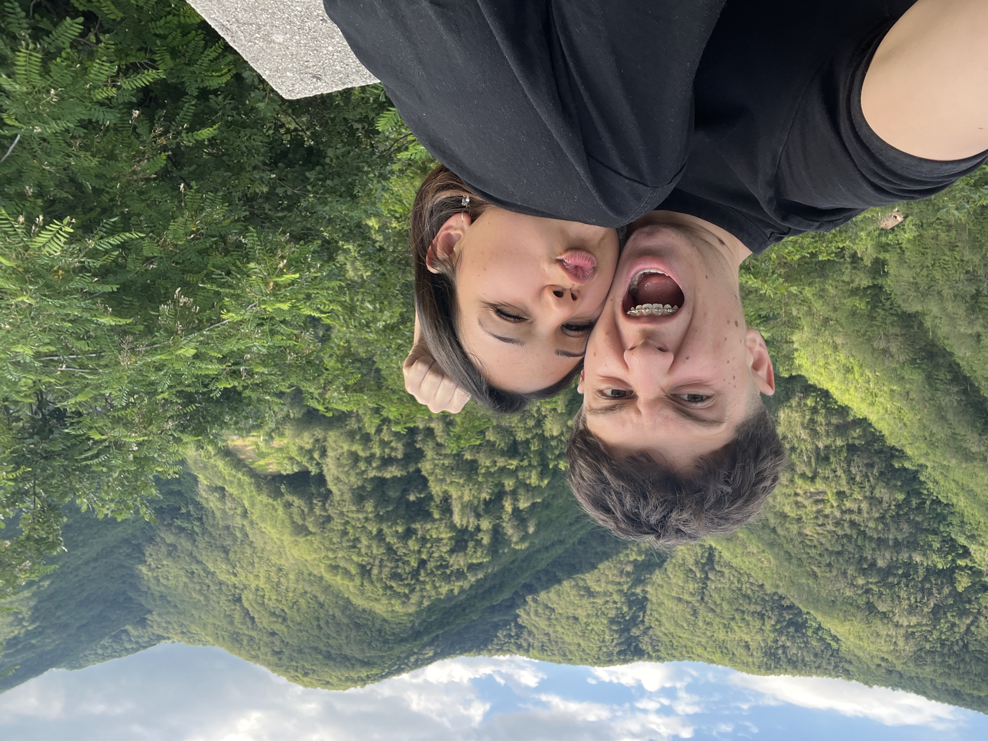 A young couple is taking a selfie with a mountainous, forested landscape in the background. The male is in the foreground, his mouth wide open in a grimace and showing his braces. The female is to his right, leaning into him with her lips puckered as if to kiss him. Both are wearing black shirts. They are standing on a stone wall. The mountains behind them are covered in lush green trees and under a partly cloudy sky. The location is the city of Pechinska, Bulgaria.