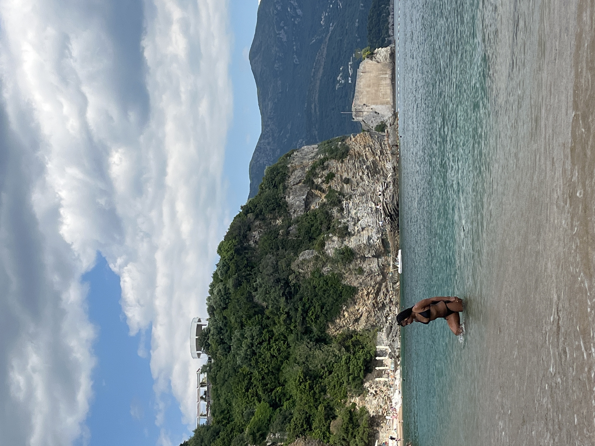 The image depicts a beach scene in Budva, Montenegro. A woman wearing a black bikini is standing in the water along the shoreline. The water is clear and turquoise in color.
Behind the beach, there is a rocky cliff covered with green trees and vegetation. A white building structure is visible on the cliff's edge. In the background, there is a taller mountain range partially obscured by clouds.
The sky overhead is filled with a mix of light and dark clouds. The overall light suggests it is daytime.