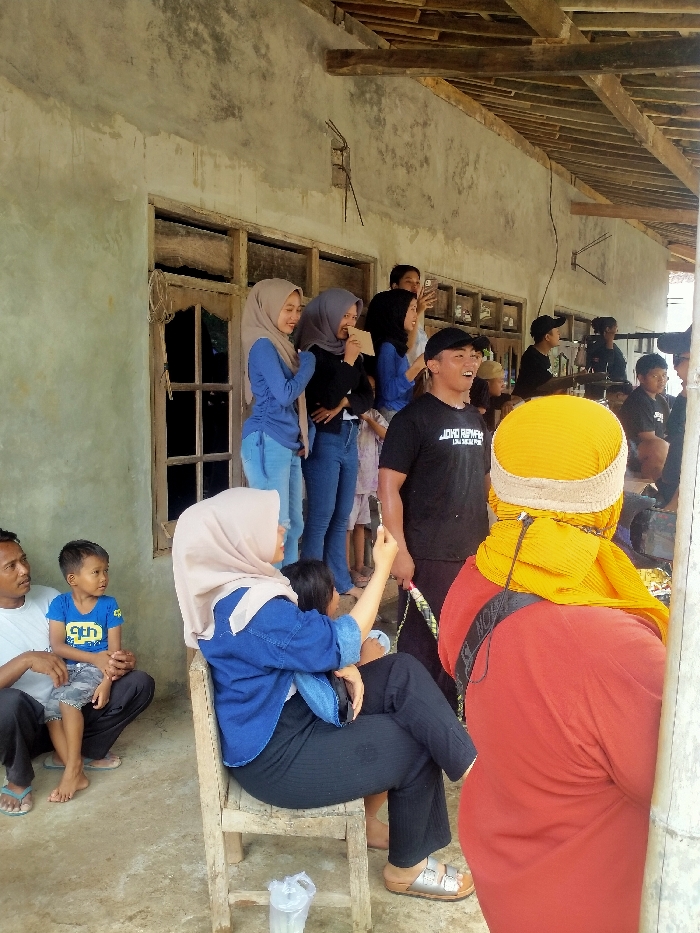 An outdoor scene in Gebyog, Indonesia, depicts a gathering of approximately fifteen individuals engaged in observation and social interaction.
In the foreground, a person in a red shirt and yellow head covering is seen from behind, facing right. To their left, a woman wearing a beige hijab, blue jacket, and black pants sits on a wooden chair, holding a child on her lap. Her right arm is raised, holding an object, possibly a mobile phone. A clear plastic cup with a white lid rests on the ground near her feet.
To the left of the seated woman, a man in a white t-shirt and dark pants sits on the ground, holding a young boy in a blue t-shirt on his lap. Both are looking towards the right.
In the mid-ground, a man in a black t-shirt (partially reading "JOKO REW") and a black baseball cap stands, facing right, with an open-mouthed expression, possibly laughing or speaking. He holds a multi-colored stick-like object.
Behind these individuals, against a weathered, light-colored wall with exposed rebar, several women are standing. Three women in the center, wearing hijabs (beige, grey, and black respectively), are smiling and looking towards the right. One holds a light-colored rectangular object. A fourth woman in a blue top and black hijab is partially visible, also looking right. Further back, several other individuals are partially visible, some also looking right, including a man in a black cap.
An open wooden window frame is visible in the wall behind the standing women. Above, a simple wooden beam structure forms an overhang or roof support. The ground is a light-colored, uneven surface. The overall atmosphere suggests an informal social event, with many participants focused on an unseen activity to the right of the frame.