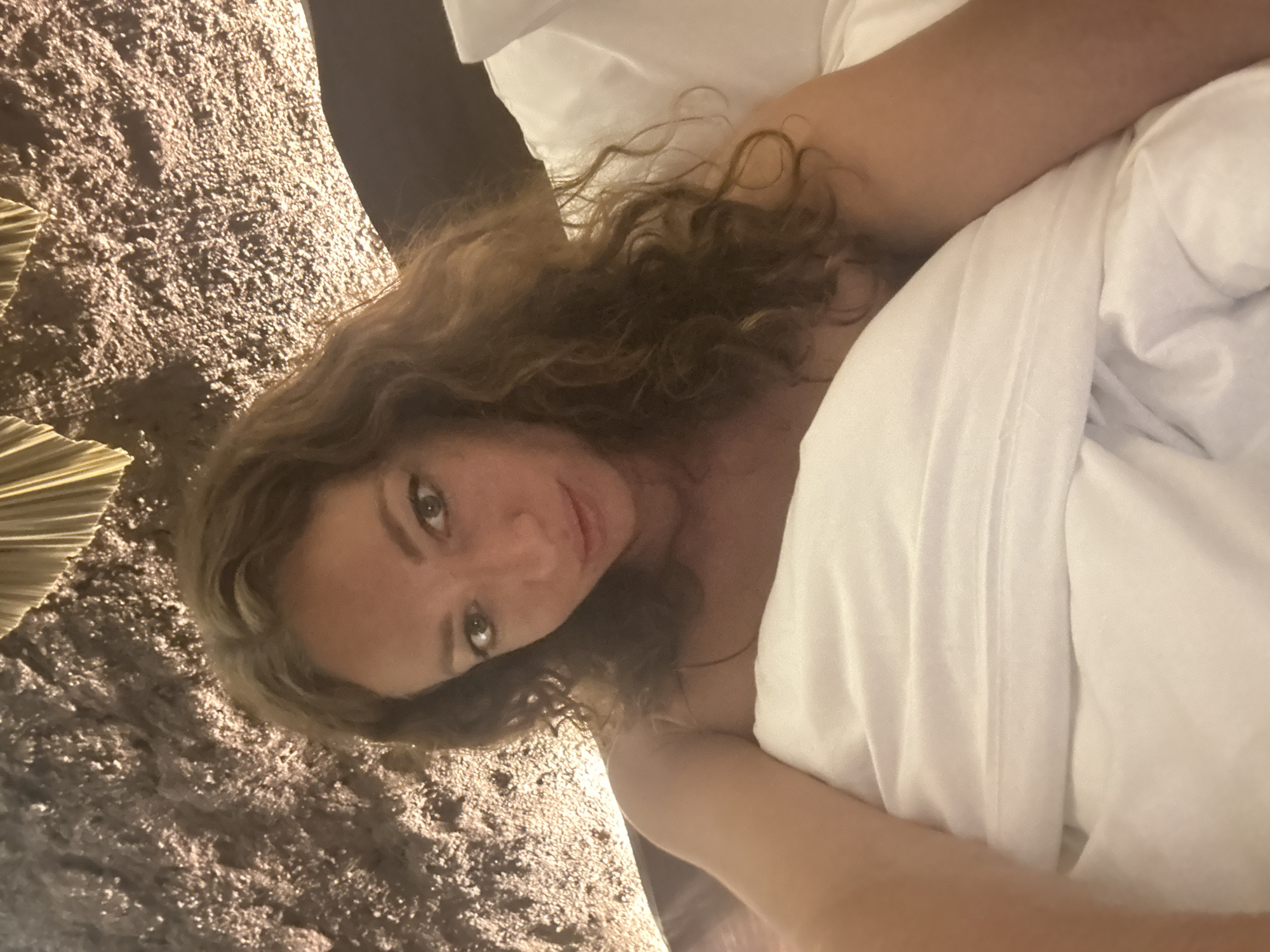 The image shows a close-up, selfie-style shot of a woman lying in bed, covered by white sheets and blankets. She has long, wavy brown hair and is looking at the camera with a relaxed expression. The background features a textured wall with warm lighting, giving a sense of an indoor, likely residential or hotel, setting. The image does not provide any specific information that would allow for its localization to Nam Hoa Lư Ward, Vietnam.