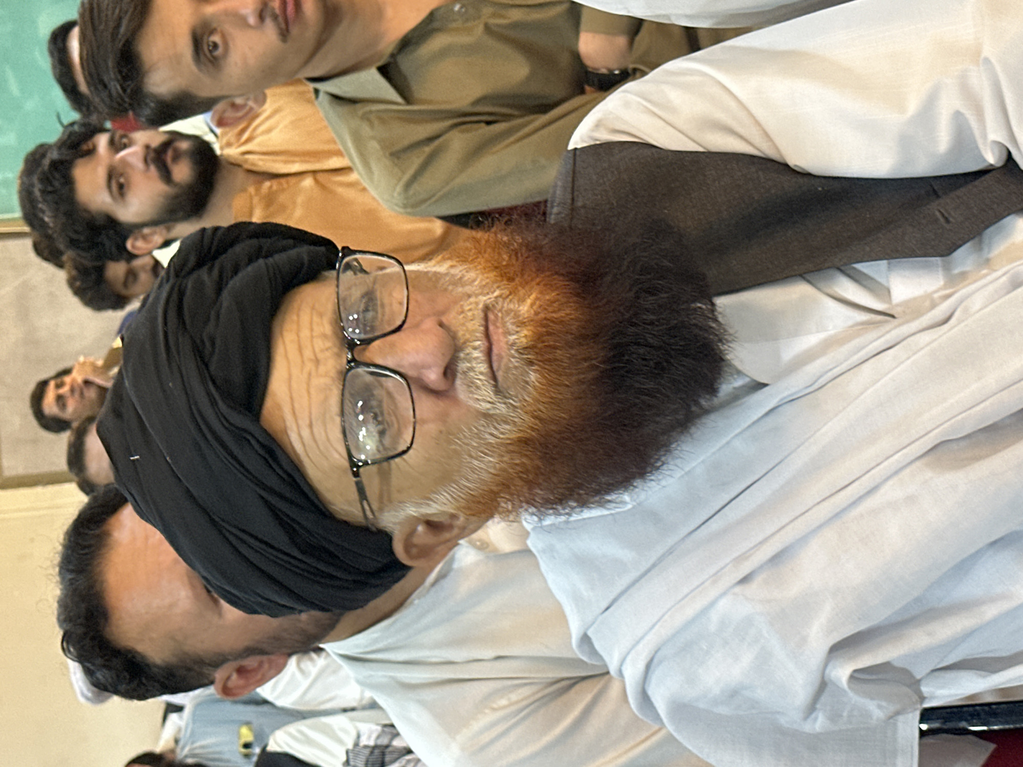 A medium shot captures an elderly man with a prominent reddish-brown beard and a black turban centered in the frame. He wears eyeglasses and a light-colored garment, with a dark vest over it. Several other men are visible in the background, some appearing younger, all dressed in traditional attire. The setting appears to be indoors, with neutral-colored walls and a hint of natural light. The context suggests a gathering or event within Quetta City Tehsil, Pakistan.
