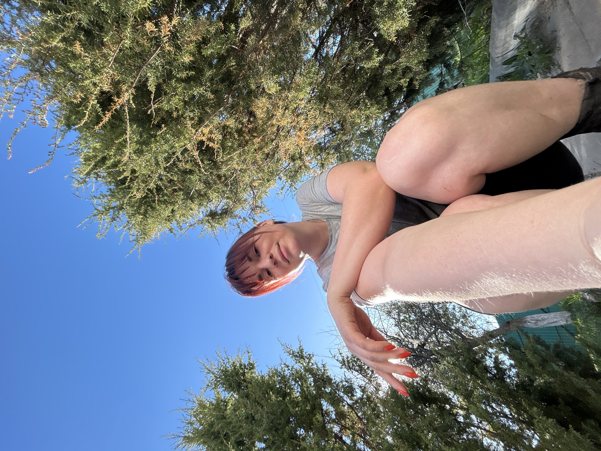 A low-angle, outdoor shot looking up at a person with pale skin, red-orange hair, and matching nail polish. The person is wearing a gray t-shirt and dark shorts, sitting with their legs tucked, arms resting on their knees. Behind the person is a clear, blue sky and dense evergreen trees with thin, needle-like leaves. In the background, some of what appears to be a structure with green siding can be seen through the branches. There is no information to support the location of the photo as the city of Фоки, Russia.