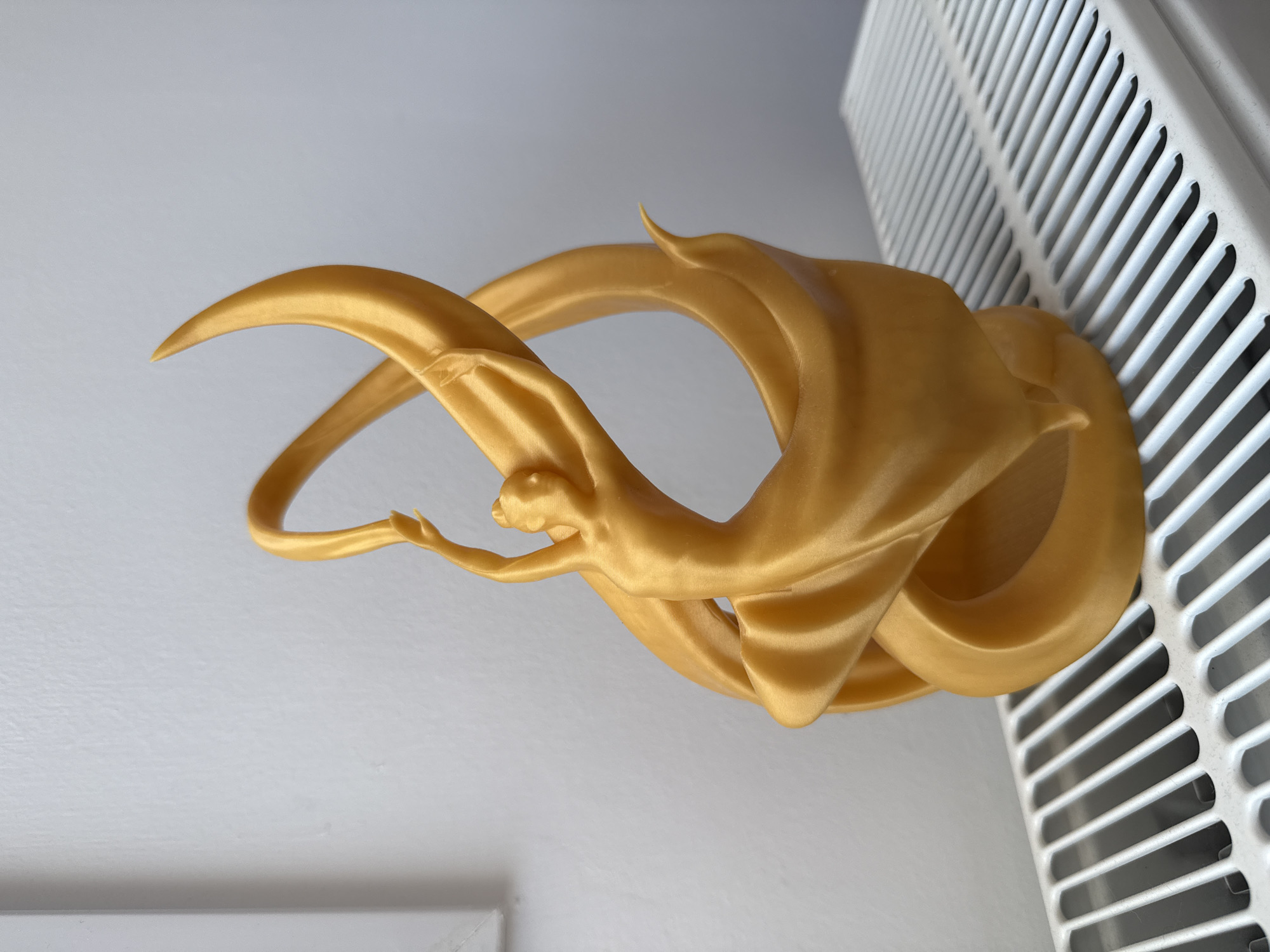 A golden, 3D-printed sculpture of a dancer in mid-leap, her arms outstretched and fabric swirling around her, sits atop a white radiator. The sculpture is illuminated from the front, casting a subtle shadow on the white wall behind it. The radiator's metal slats are clearly visible beneath the sculpture. The scene is indoors, with a plain white wall as the backdrop. No people or animals are present in the image. The location is identified as Neoi Epivates, Greece.