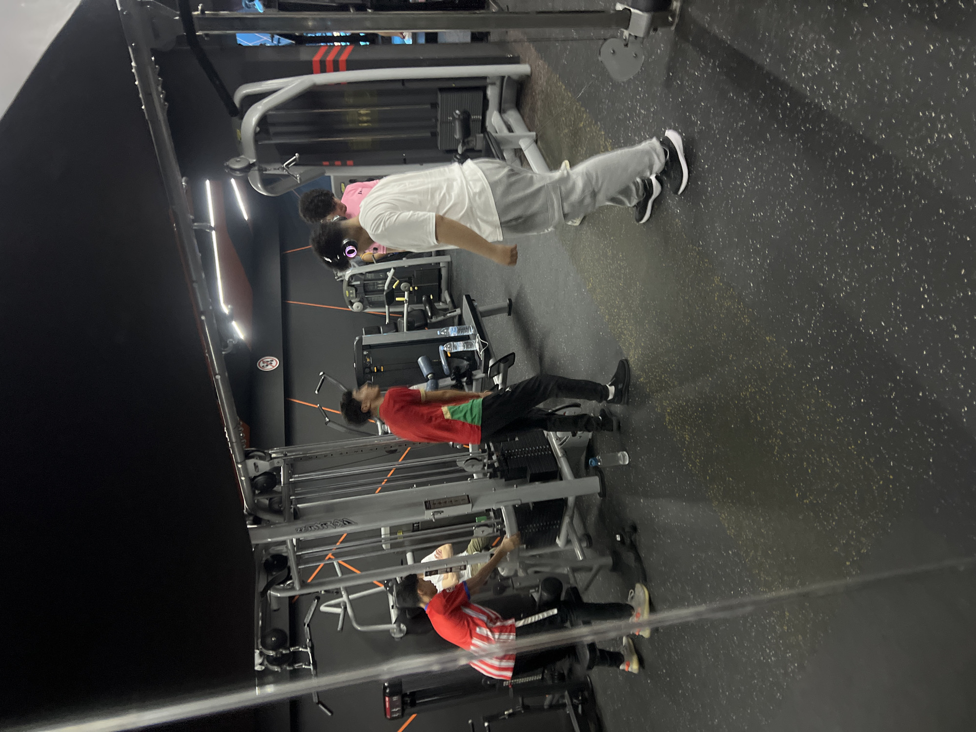The image captures an indoor gym scene in M'Diq, Morocco, seemingly taken through a reflective surface like a mirror or glass, causing a layered and slightly blurred effect in the foreground. Several young men are present, engaged in or preparing for exercise.
In the most prominent position, a young man with dark, curly hair is seen wearing a white t-shirt, grey sweatpants, black and white sneakers, and over-ear headphones. He stands upright, facing towards the right side of the frame, with his arms slightly bent, appearing to be resting or observing. His reflection is faintly visible in the lower part of the foreground.
Further back, another young man, also with dark hair, wears a red t-shirt with a green accent on the side, dark trousers, and black sneakers. He stands near a cable machine, appearing to be focused on the equipment.
In the background, a third individual is visible, wearing a red and white striped t-shirt and dark pants. This person is actively interacting with a piece of gym equipment, possibly a cable machine, suggesting they are in the process of exercising.
The gym features dark grey or black walls, some with three prominent red horizontal stripes, and a dark, speckled rubber flooring. Various pieces of modern gym equipment, including multiple cable machines and weight stacks, are arranged throughout the space. Artificial lighting illuminates the area. A small, circular logo, possibly "CR" or similar text, is visible on one of the black walls. The setting suggests a casual, active training environment.