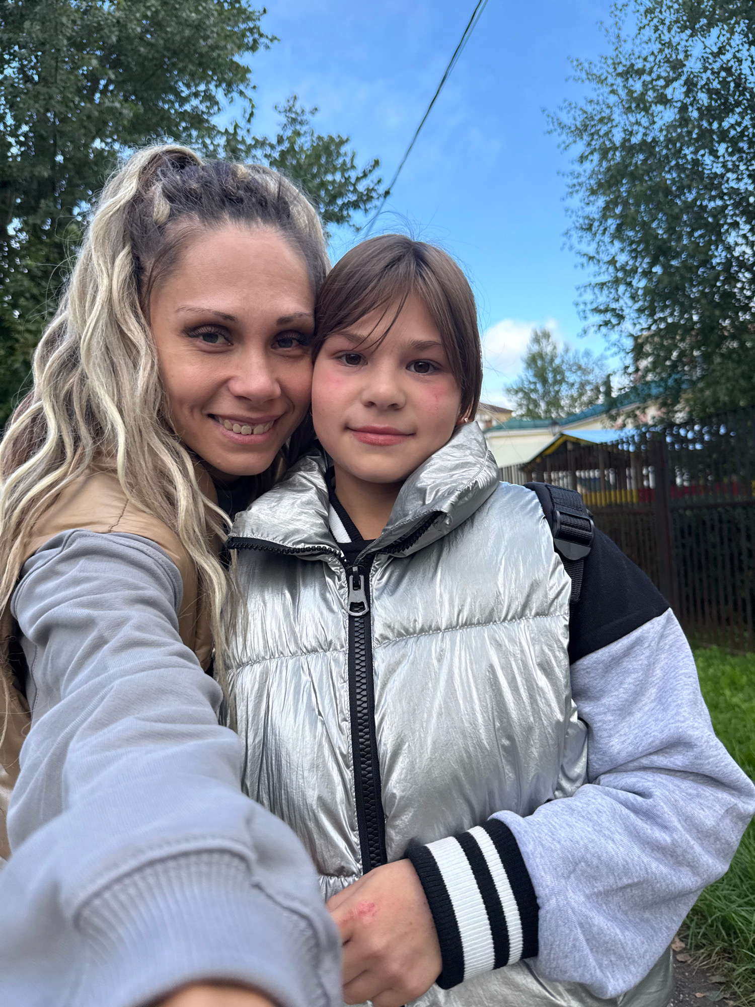 In this photograph, a woman and a child are captured in a selfie outdoors. The woman, with blonde hair styled in a high ponytail, smiles at the camera. The child, positioned next to her, also looks towards the lens. Both are wearing casual outdoor clothing. The background reveals trees and a blue sky with scattered clouds, suggesting a daytime setting. In the distance, a fence and a building are visible, indicating an urban or suburban environment. The presence of a fence and a building in the background, coupled with the vegetation and sky, aligns with the characteristics of a location like Rusanovo, Russia.