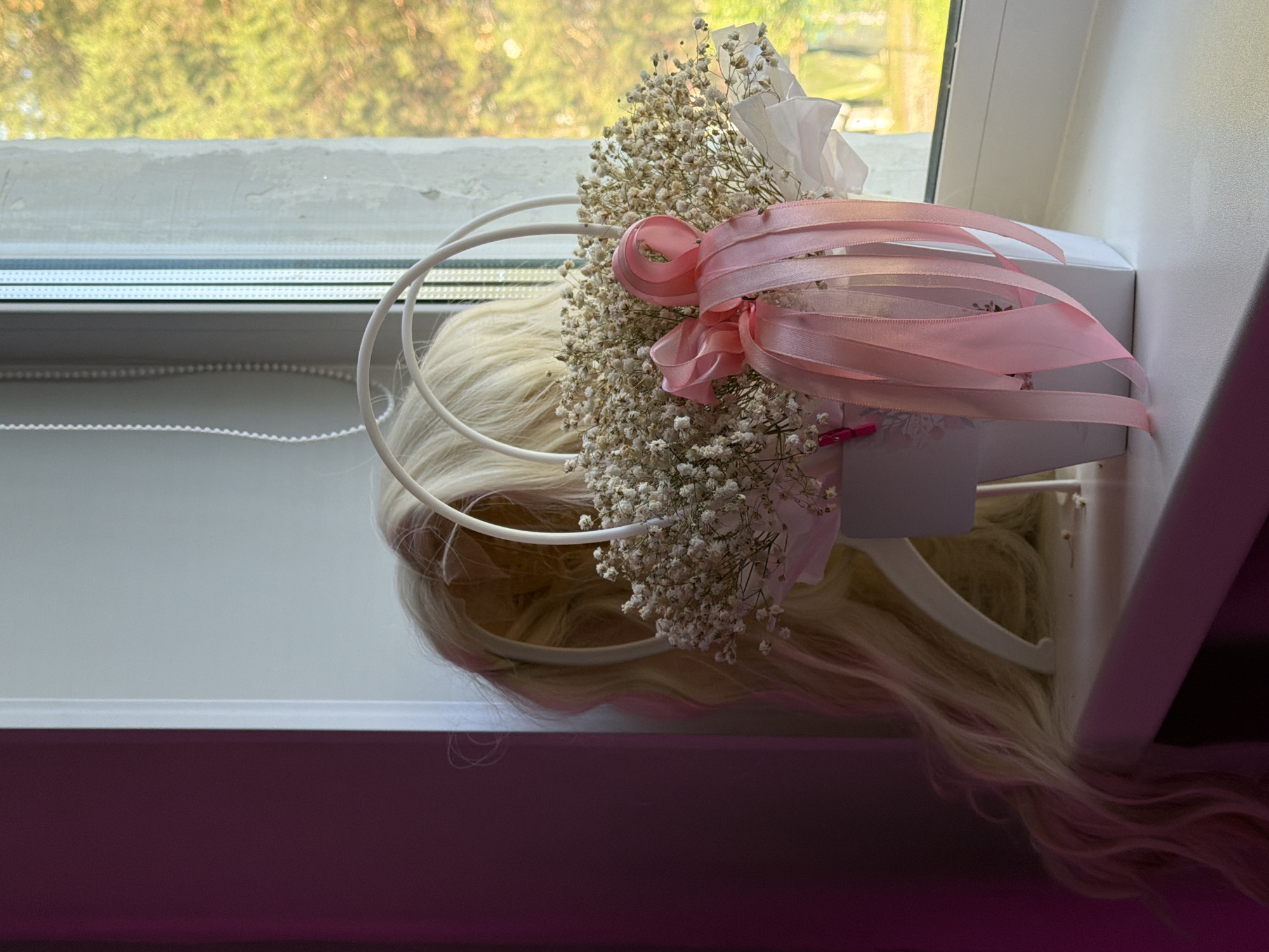 A blonde wig is placed in a white basket with a pink bow and ribbons. The basket contains a bouquet of dried baby's breath flowers. The wig and basket are on a windowsill. To the left is a dark purple wall, and to the right is a window with a view of trees. The scene is indoors, and the lighting suggests it is daytime.