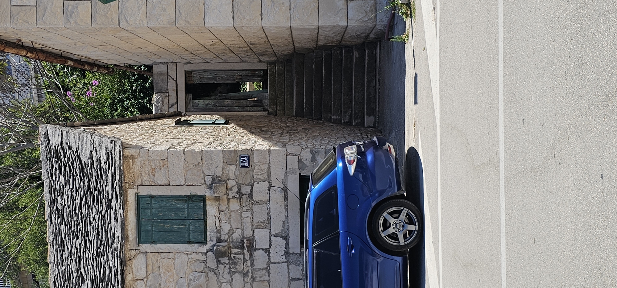 A blue station wagon is parked on an asphalt road in front of a stone building with a slate roof. The building has a green shuttered window and an open wooden door leading to a set of stone stairs. To the right of the stairs is another stone building. The scene is set in Općina Pučišća, Croatia, with lush green trees and vegetation in the background.