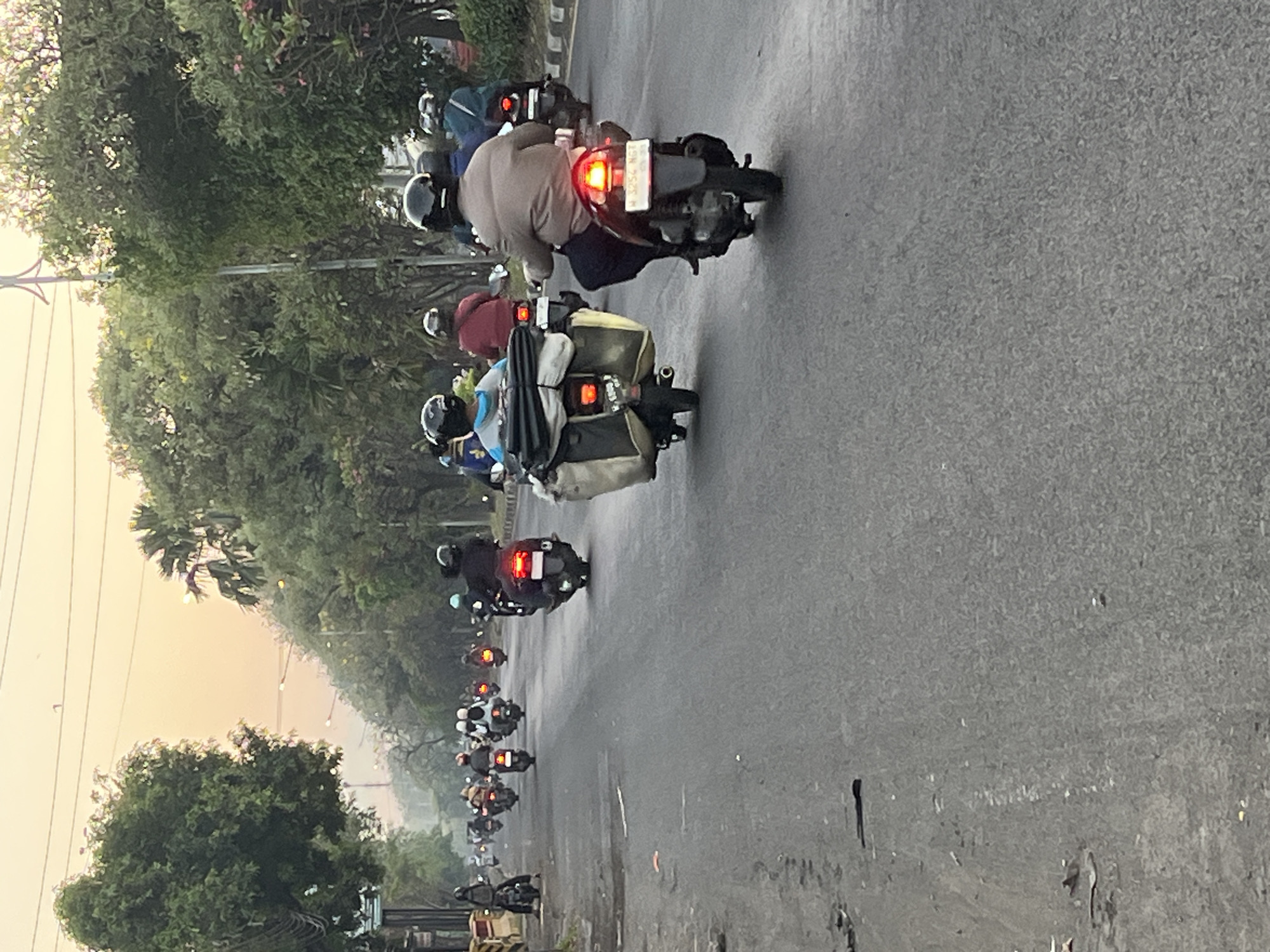 The image depicts a street scene in Ngaban, Indonesia, with a convoy of motorcycles traveling along a paved road lined with trees and vegetation. The time of day appears to be late afternoon or early morning, as indicated by the soft, warm lighting and the overcast sky.
The foreground is dominated by the road, which shows signs of wear and tear, with some patches and debris visible. Several motorcycles are in motion, with riders wearing helmets. The motorcycles are of various models, and some are carrying cargo, such as bags attached to the rear.
In the middle ground, more motorcycles are visible, forming a procession. The riders are primarily wearing casual clothing. The surrounding environment features dense foliage and trees, suggesting a tropical or subtropical setting. Power lines are also visible overhead.
The background reveals a continuation of the road and more trees. The overall impression is of daily life and transportation in a bustling urban or semi-urban environment in Indonesia.
